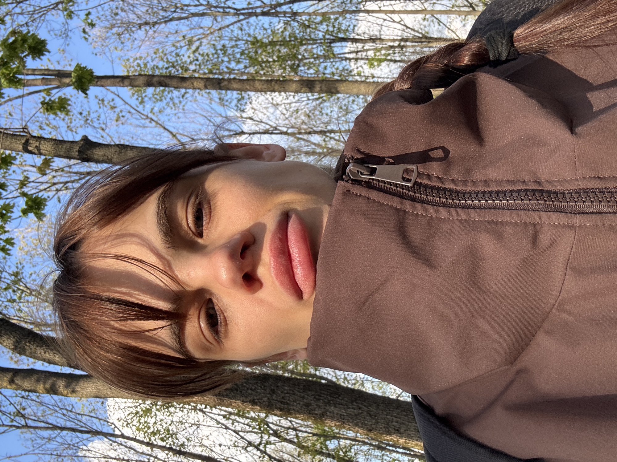 A close-up, rotated portrait shot captures a young person, likely female, taking a selfie outdoors. The subject's face is prominent on the left side of the frame, featuring brown hair with bangs that fall across the forehead. Her eyes are open, and her lips are slightly parted in a pout. Strong sunlight illuminates her face, highlighting her features and casting subtle shadows.
She is wearing a brown jacket with a visible zipper and pull-tab on the right side of the image. A black scrunchie holds her brown hair in a braid, partially visible at the top right, indicating the hair is pulled back.
The background reveals a clear, bright blue sky dotted with white clouds. Numerous bare or sparsely leafed tree branches and trunks stretch across the upper and central parts of the background, suggesting a time of year like early spring, where leaves are just beginning to emerge. The trees appear tall, implying a park or natural outdoor setting in Rostov-on-Don, Russia. The overall impression is a sunny, daytime scene with a relaxed atmosphere. No text is visible in the image.