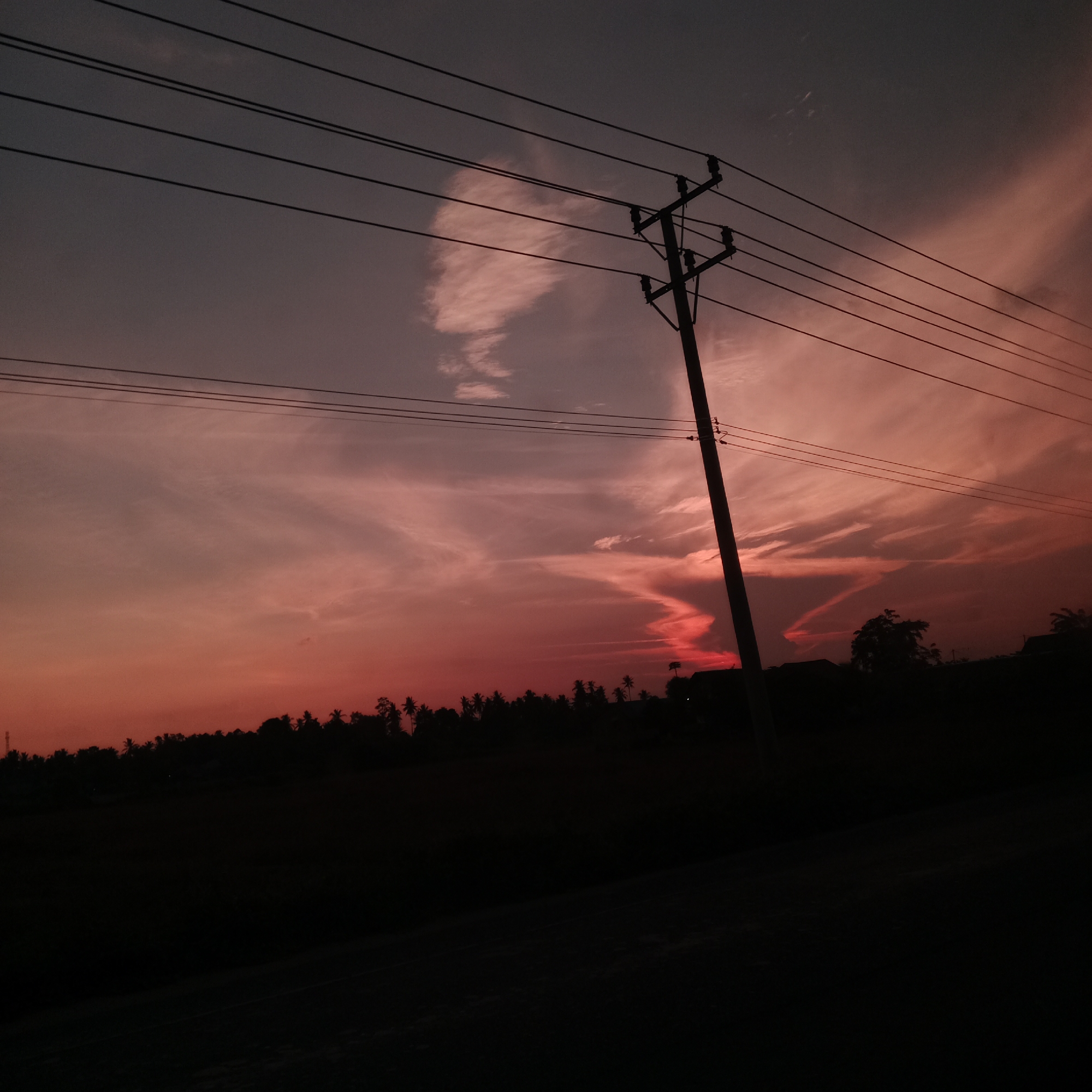 The image depicts a silhouetted landscape at sunset, viewed from the perspective of Cotabato, Indonesia. A prominent power pole stands on the right side of the frame, with multiple electrical wires stretching across the upper portion of the image. The sky is a gradient of deep purple and vibrant orange, with wispy clouds catching the fading sunlight. In the background, a dark, dense treeline, likely comprised of palm trees characteristic of the region, forms the horizon. The foreground is a black silhouette of the ground, possibly a road or field. There are no people or visible actions taking place. The overall atmosphere is tranquil and captures the serene beauty of a tropical sunset.