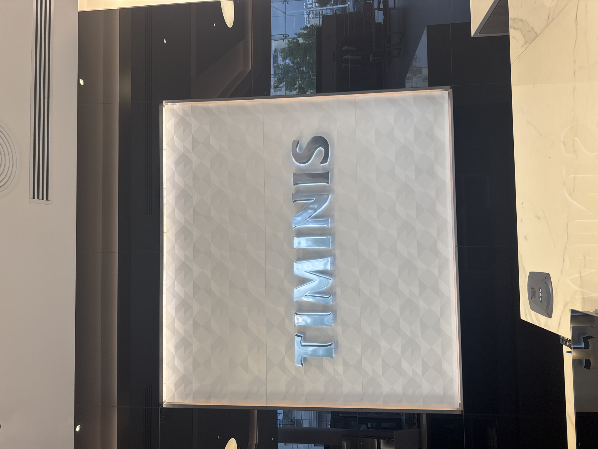A wide shot shows the interior of a commercial establishment, likely a high-end store or showroom, in Limassol, Cyprus. The main focal point is a large, illuminated sign that reads "TIMINIS" in large, three-dimensional metallic letters. The letters are positioned against a textured, geometric white background with a subtle diamond pattern, illuminated by soft, diffused light from above. The overall aesthetic is modern and sophisticated.
To the left of the sign, a dark, reflective surface, possibly a large mirror or glass panel, shows a partial reflection of the interior and the exterior environment. Through this reflective surface, one can discern elements of an urban landscape outside, including buildings and trees, indicating the store is situated in a city.
Above the sign, a dark band of paneling runs across the width of the scene, with a linear air vent and a circular ceiling fixture visible. Below the sign, a dark, solid base supports the illuminated display. Further down, in the foreground, a portion of a sleek, marble-topped counter or display area is visible. On this surface, a dark gray electrical outlet or power hub is present, along with a sliver of a metallic object, possibly part of a fixture or stand. The lighting in the foreground is dimmer, with ambient light reflecting off the marble. The scene appears to be devoid of any visible people.