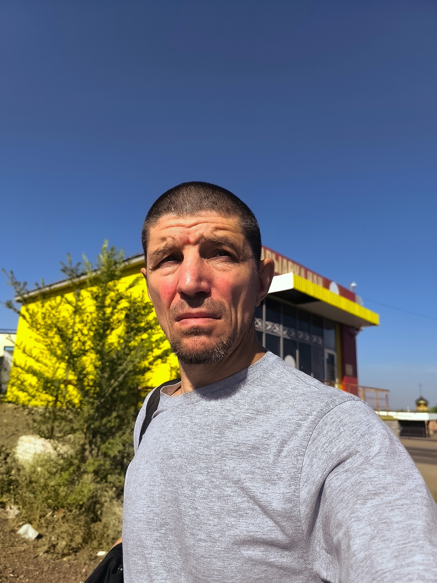 A man is captured in a selfie outdoors, with a bright blue sky as the backdrop. He has a short haircut, stubble, and appears to be in his late 30s or 40s. His expression is serious, with his brow furrowed. He is wearing a grey t-shirt, and a black bag strap is visible over his shoulder.
Behind him, a vibrant yellow building with a red and yellow striped awning stands out. A leafy green tree is positioned in front of the building, adding a splash of natural color to the scene. To the far right, a golden dome, possibly belonging to a religious building, is visible against the horizon, suggesting a potential location clue. The sun is shining brightly, casting strong shadows and highlighting the textures on the man's face and clothing, indicating it is daytime.
The overall impression is of a candid moment captured on a sunny day, possibly in an urban or semi-urban environment given the building and the hint of the dome. There is no visible text in the image.