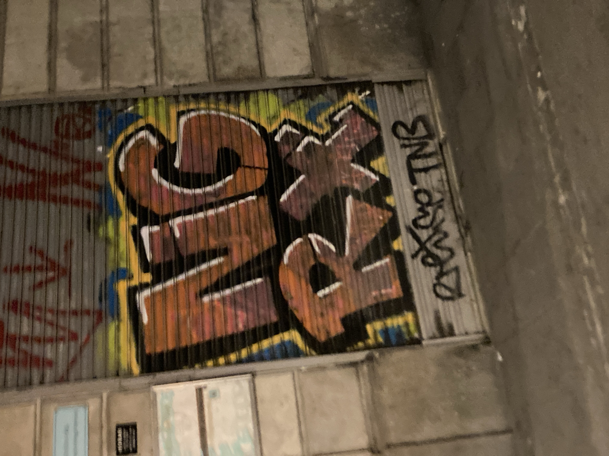 The image depicts a close-up view of a graffiti-covered metal roll-up door in an urban setting, likely a shop or business. The graffiti features large, bold lettering in orange and pink tones, with a black outline and white highlights. Below the main lettering, there is a smaller tag in black. The door is installed on a building with a textured facade, possibly made of concrete blocks, on the right side of the image. To the left of the door, there are sections of a light-colored wall with some signage and what appears to be a partially visible map or poster. The street in front of the door is visible in the lower part of the image, showing a paved surface. The lighting suggests it is nighttime or a dimly lit area, with some ambient light illuminating the scene. The graffiti appears to be the primary subject of the photograph, highlighting a form of street art on a utilitarian urban surface. Given the style of architecture and the presence of signage, it is plausible this scene is located in a commercial area of a European city. Without more specific visual cues in the graffiti or signage, pinpointing the exact location within Barcelona is not possible from this image alone.