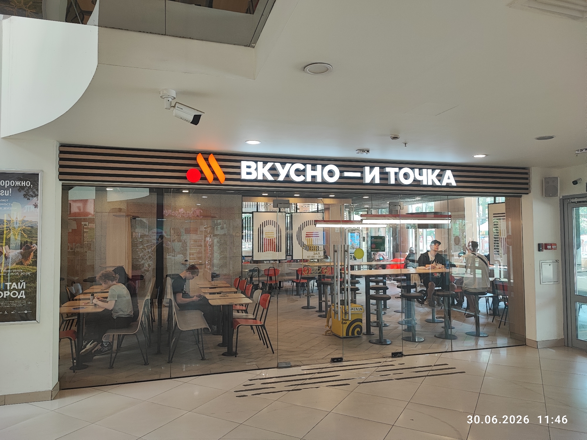 The image shows the interior of a fast-food restaurant named "ВКУСНО — И ТОЧКА" (Vkusno — i Tochka), which is a Russian fast-food chain that replaced McDonald's. The restaurant has a modern design with large glass windows allowing a view into the dining area and the kitchen. Inside, there are several tables and chairs for customers, some of whom are seated and appear to be eating or using their phones. The lighting is bright, typical of a commercial establishment.
On the left side of the image, a poster is displayed on the wall. The restaurant's signage is prominently visible above the entrance. The floor is tiled, and there are some decorative elements on the walls. The overall impression is of a clean and well-lit dining space.
The timestamp "30.06.2026 11:46" is visible in the bottom right corner of the image, indicating the date and time the photo was taken. There are no visible indications that this scene is specifically located in Lytkarino, Russia.