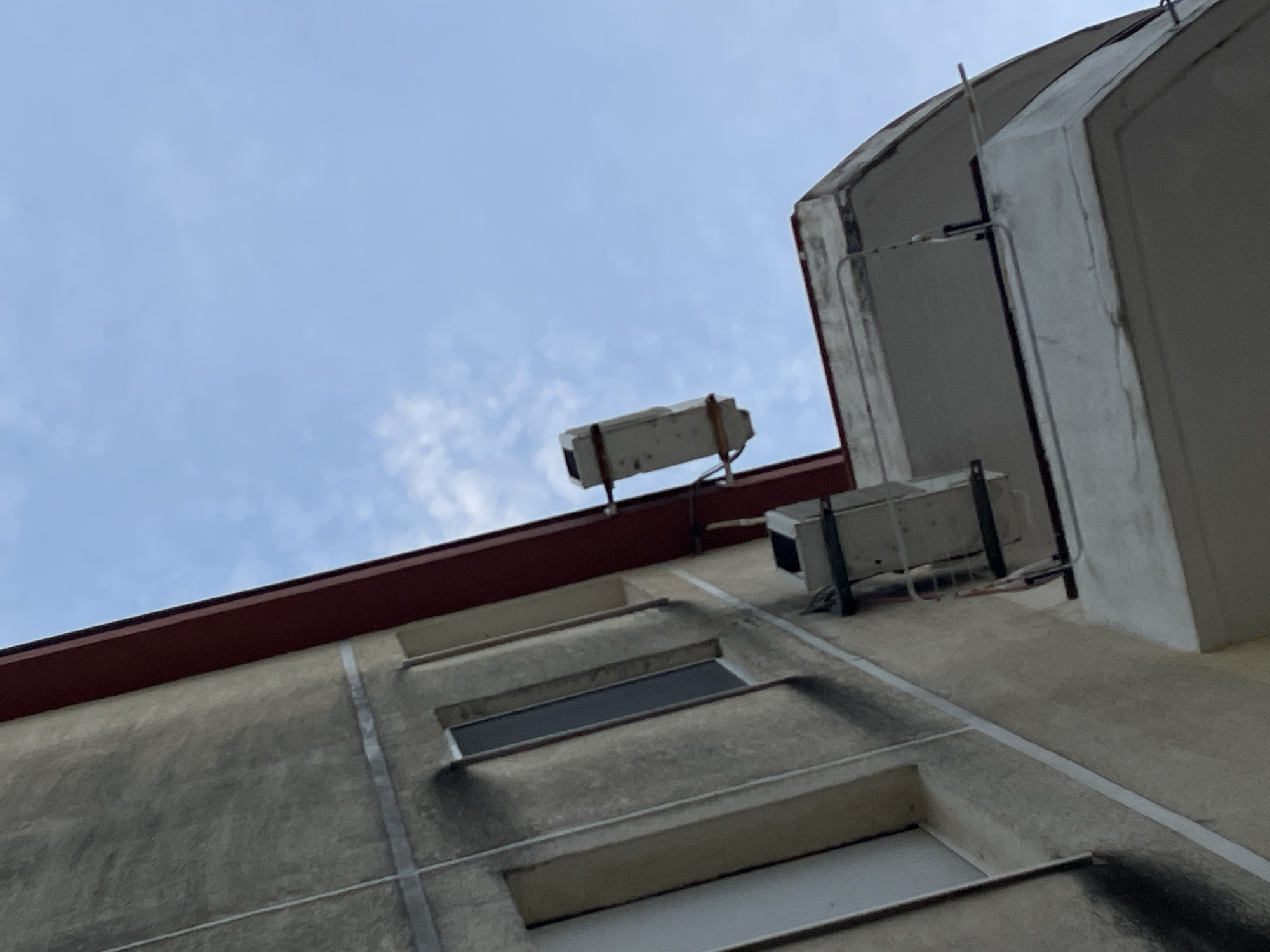 A low-angle shot captures a section of a weathered, grey concrete building under a pale blue sky with scattered clouds. The building features a red roofline and evenly spaced rectangular windows, some of which appear darkened. Mounted on the building's roof and upper wall are two rectangular, white objects that appear to be electrical boxes. The overall environment suggests an urban setting, likely in Budva, Montenegro, given the building's architecture and context.
