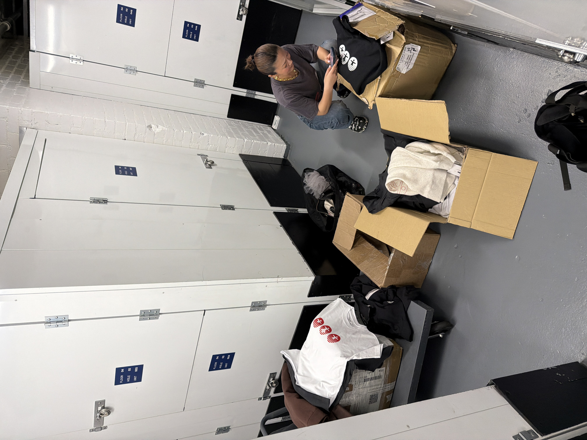 The image shows a woman taking pictures of products in a storage unit in New York City. The units are white, with labels indicating floor, aisle, and unit numbers. The woman is kneeling and holding a smartphone to photograph a black tote bag in a cardboard box. Other cardboard boxes are scattered around the unit, containing clothing items. The floor is grey concrete, and the lighting is bright and somewhat sterile.