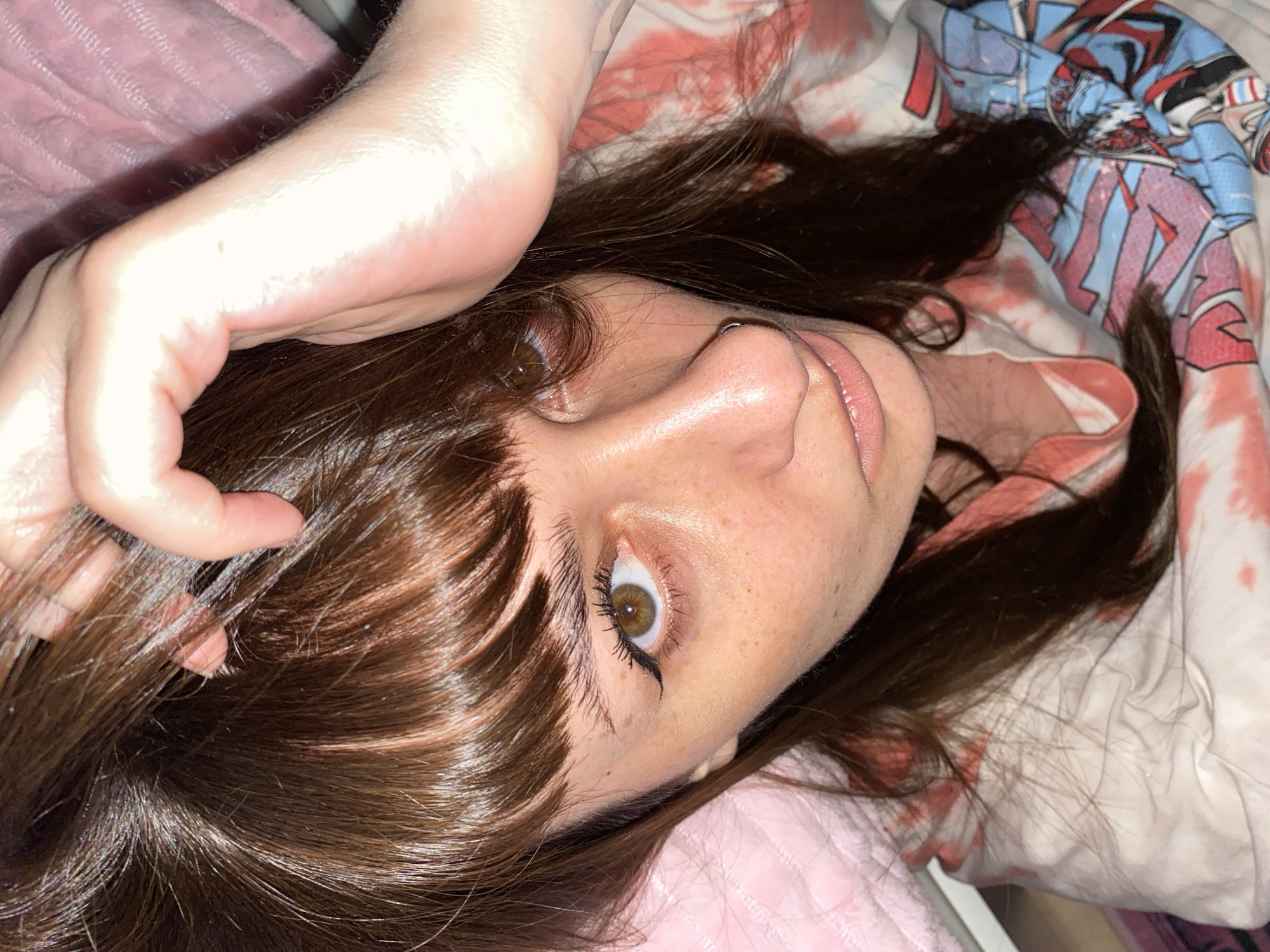 The image presents a close-up, high-angle portrait of a young individual lying down, looking directly at the camera. The setting appears to be indoors, likely a bedroom in Votkinsk, Russia, based on the provided context and the visible bedding.
The subject has long, dark brown, shiny hair with bangs that partially cover their forehead. Their left arm is raised, with the hand resting on or near their head, pushing some hair back. They have fair skin with noticeable freckles across their nose and cheeks. Their wide-open eyes are hazel or light brown, emphasized with black winged eyeliner and mascara. A small, silver septum nose ring is visible. Their lips are slightly parted, showcasing a natural pink color.
The person is resting on or surrounded by various textiles. A pink, textured blanket or comforter is prominent on the left and bottom of the frame. In the upper right, there is another piece of fabric, possibly a pillowcase or blanket, featuring an abstract pattern of red, white, and blue shapes on a white background, which could be stylized characters or designs. The artificial lighting creates a warm glow, highlighting the sheen on the subject's skin and hair. The overall impression is a relaxed and candid portrait. No visible text is present in the image.