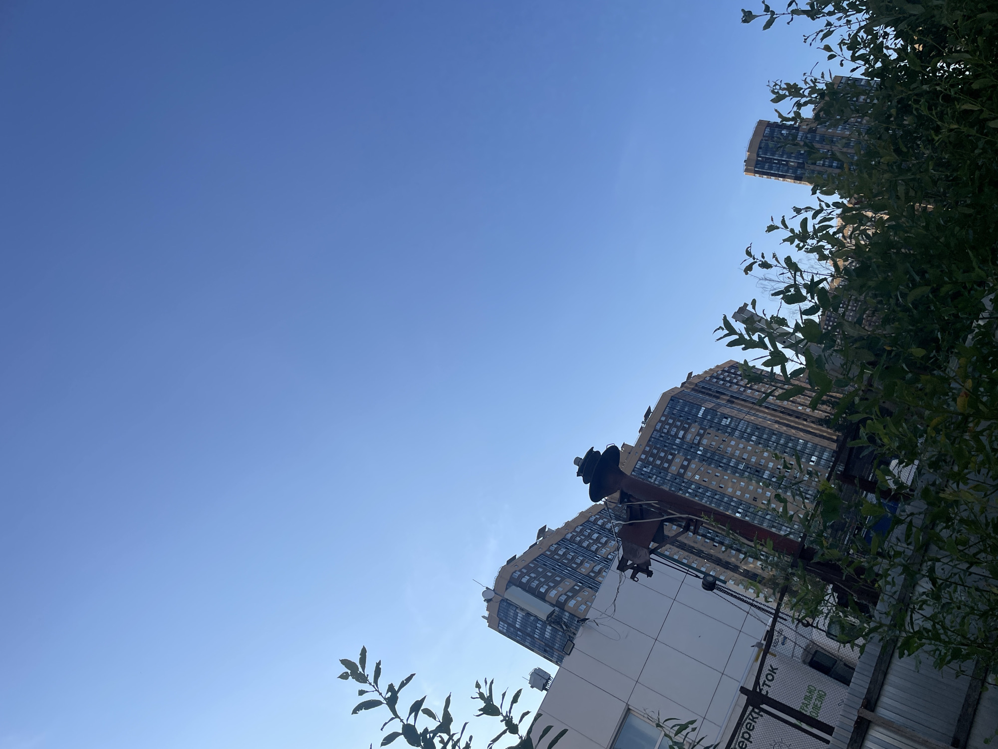 In this low-angle shot of a construction site in an unknown city in Russia, a bright blue sky dominates the upper half of the frame. In the lower portion, several tall apartment buildings are visible, their facades characterized by a grid of windows and varying shades of beige and brown. In the foreground, lush green foliage obscures a portion of the scene, suggesting the presence of trees or bushes. A metal fence runs along the bottom of the image. To the left, a section of a white building with a window is visible, bearing Cyrillic text which translates to "PEREKRYSTOK" and "PRAVILNO POLEZNO" (correctly useful). Amidst the greenery and structures, a large industrial crane stands prominently, its rusty metal frame a stark contrast to the newer buildings and the clear sky. The scene appears to be set during daylight hours, with the sun likely positioned to the left, casting light on the buildings.