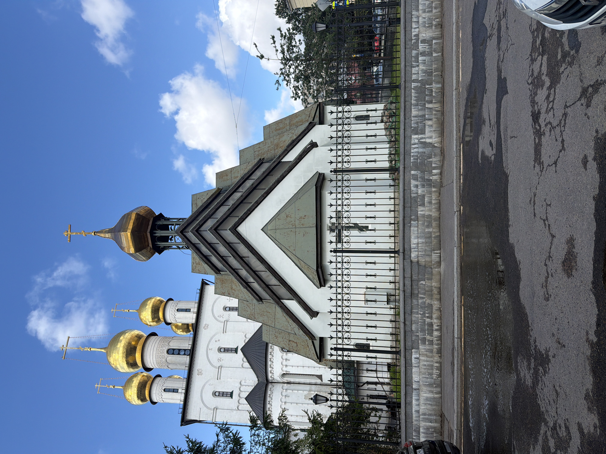 A white church with golden domes is seen under a partly cloudy blue sky in Saint Petersburg, Russia. The church features multiple domes, with the largest centrally located and adorned with a gold cross. The building exhibits both traditional and modern architectural elements, with a white facade decorated with arched windows and decorative elements, and a newer section with a multi-faceted roof. A black wrought-iron fence encloses the church grounds, with a low stone wall at its base. The street in front of the church is wet and reflective, indicating recent rain, with puddles and patches of water. Trees are visible in the background, along with other buildings and a street lamp.