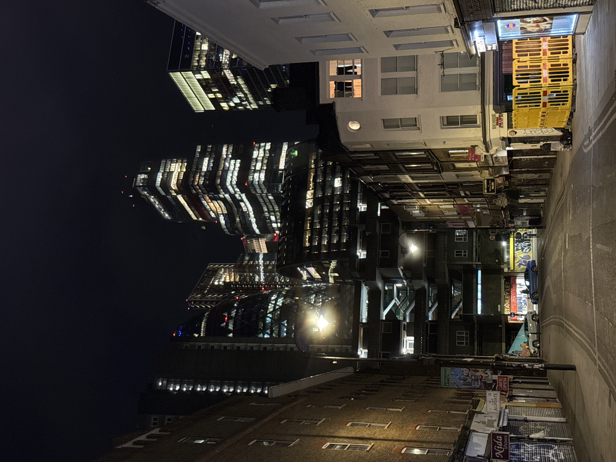 The image depicts an urban street scene at night in London, United Kingdom. Tall modern skyscrapers with illuminated windows loom in the background, contrasting with the older, shorter buildings lining the narrow street. The street is illuminated by streetlights and neon signs. On the right, a yellow barrier is stacked outside a store. A car is parked along the road. There are posters and graffiti on the buildings along the street. The street appears to be wet.