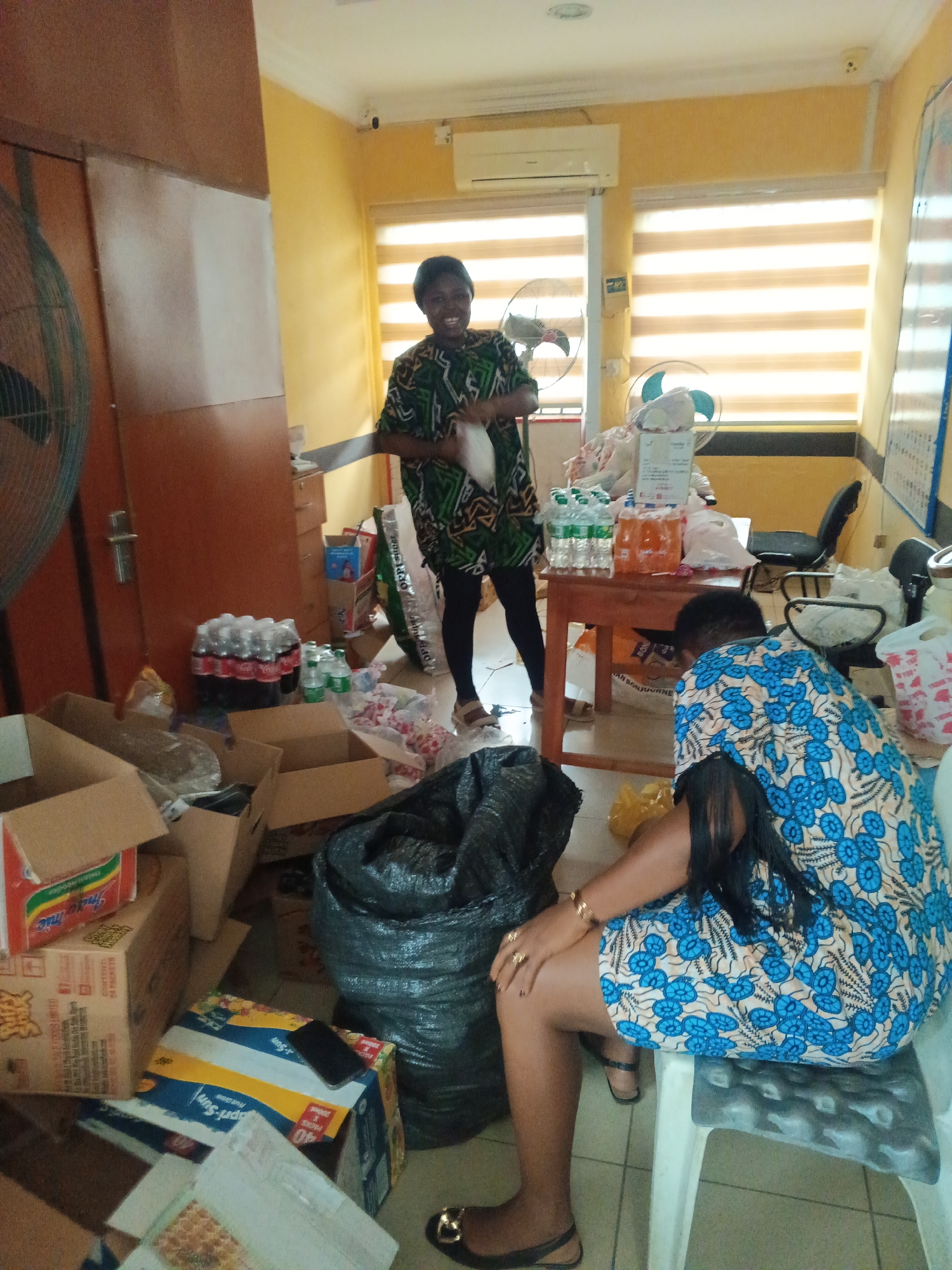 The image shows an indoor scene with two women in a room surrounded by boxes, containers of drinks, and a large black trash bag. One woman is standing, wearing a green and black patterned dress with black leggings, and appears to be holding a white plastic bag. The second woman is seated on a white plastic chair, wearing a blue and white floral patterned dress.
The room appears to be cluttered, with numerous cardboard boxes, some containing instant noodles and other food items, scattered on the floor. Bottles of Coca-Cola and water are visible, along with other beverage containers on a wooden table. Two fans are present, one in the background near the windows and another in the foreground beside a tall wooden cabinet. The room has yellow walls, and the windows have horizontal blinds.
The location is unknown, but the context suggests an indoor space possibly related to storage, packing, or distribution of goods.