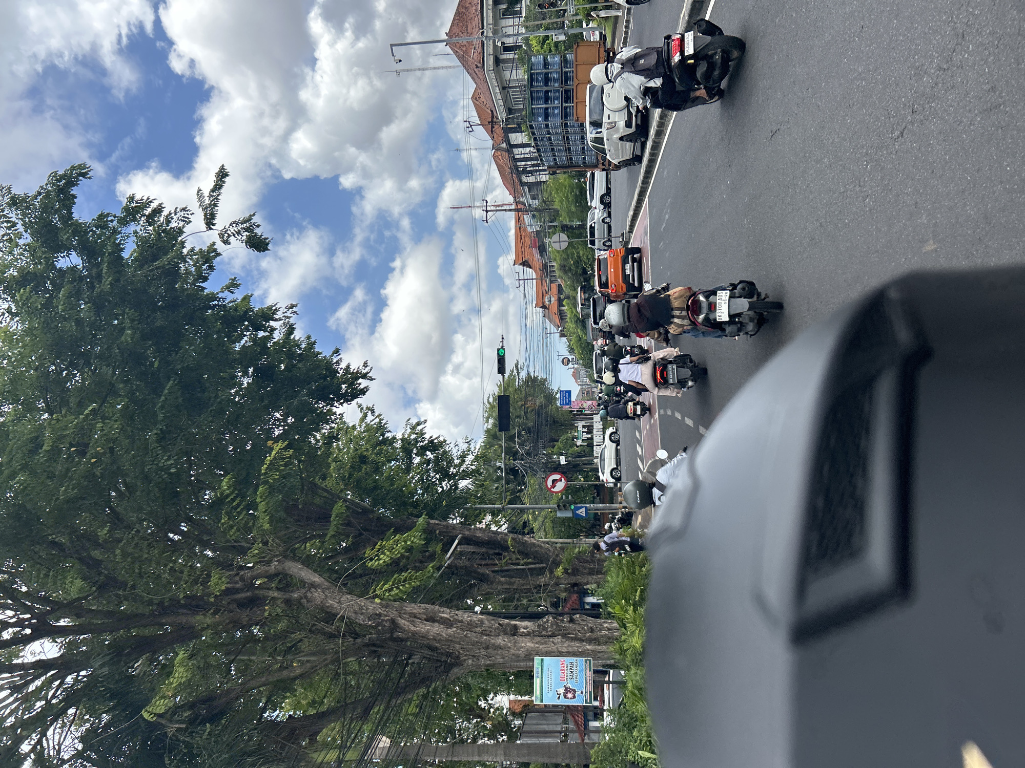 The image depicts a street scene in Denpasar, Indonesia, with a significant amount of vehicular traffic, primarily motorcycles. The perspective is from a rider on a motorcycle, looking forward. In the foreground, the dark, matte surface of a helmet visor is visible, partially obscuring the view. The road is paved, with clear lane markings. Numerous motorcycles are in motion, some carrying one rider, others with a rider and a passenger. Most individuals on the motorcycles are wearing helmets.
On the sides of the road, large, mature trees with dense green foliage line the street, casting shadows. Beyond the trees, buildings with traditional tiled roofs are visible, some appearing to be commercial establishments. A prominent white building with a dark tiled roof is situated further down the street. Traffic signals are present, with one showing a green light. There are also various signs, including advertising billboards and traffic signs, such as a circular sign with a red border and a white arrow indicating a prohibited turn.
In the background, a large truck carrying stacked blue containers is parked or moving slowly on the road. The sky is partially cloudy, with patches of blue sky interspersed with white, fluffy clouds. The overall impression is a busy urban street with typical traffic flow and architecture found in Denpasar.