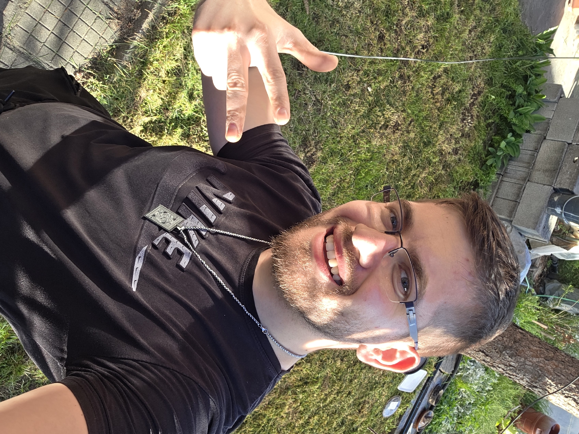 A man is taking a selfie outdoors. He is wearing glasses, a black t-shirt, and a silver chain necklace. He is smiling and holding up his hand. The background consists of green grass, some weeds, and paving stones. Part of what looks like a greenhouse can also be seen. There is no way to confirm the location is Yagodovo, Bulgaria from just the image.