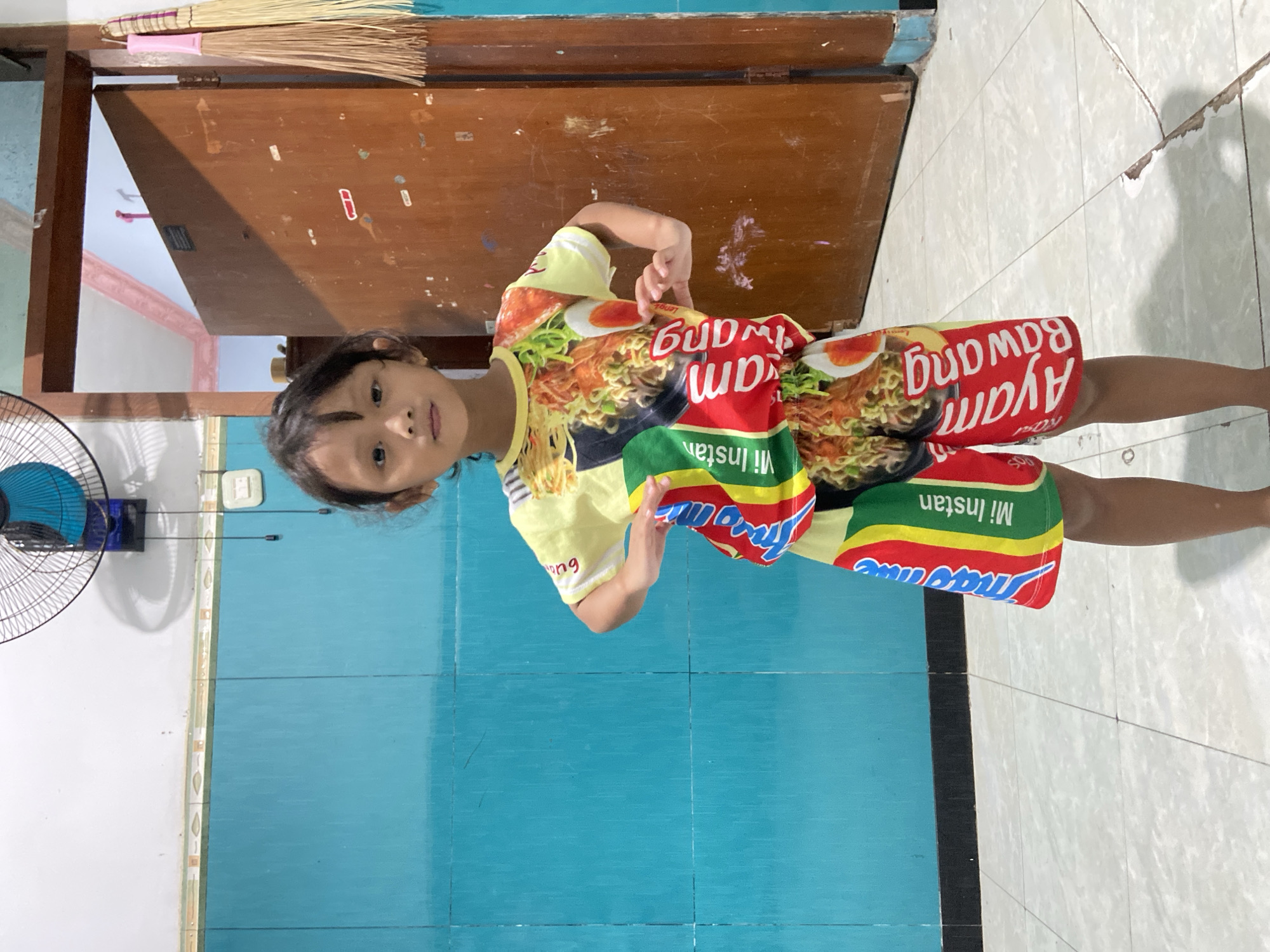 A young girl stands in a room, striking a pose with her hands on her hips. She is wearing a yellow t-shirt and shorts with a pattern resembling instant noodle packaging. The t-shirt features images of noodles, eggs, and text that appears to be in Indonesian, including "Mi Instan" and "Ayam Bawang". The shorts have a similar design. The background consists of a turquoise wall, a brown wooden door, and a tiled floor. A ceiling fan is visible in the upper left corner of the image. The lighting suggests an indoor setting. The image was taken in Gelam, Indonesia.