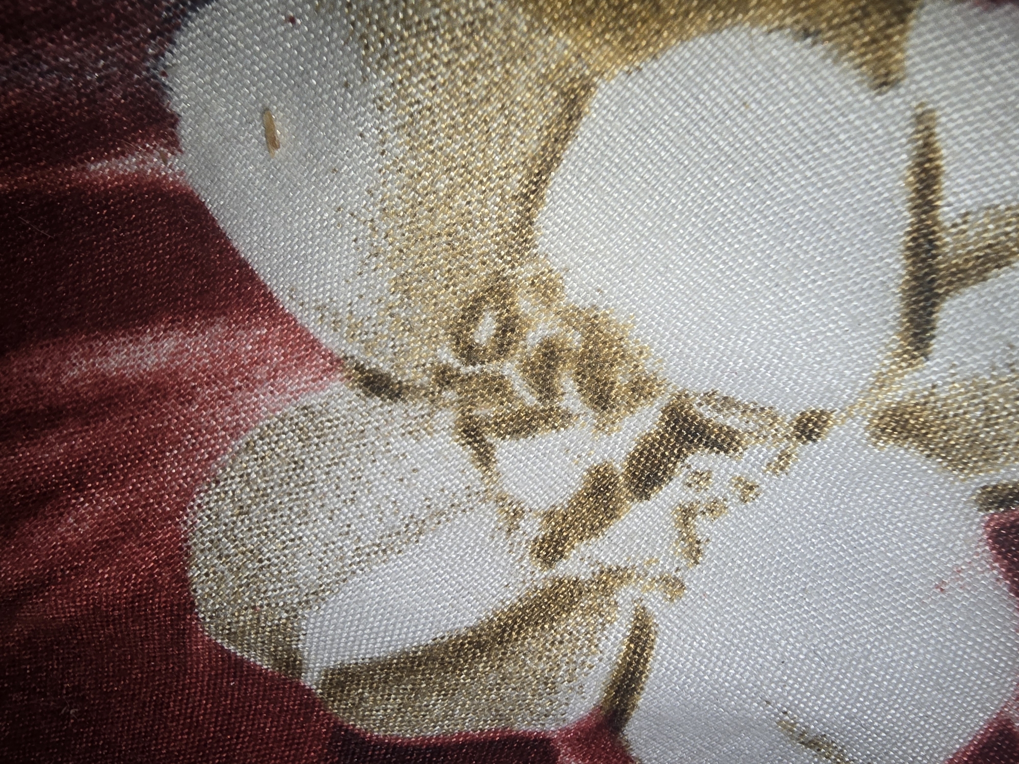 The image displays a close-up of a fabric with a floral pattern. The fabric has a textured weave, appearing to be a natural fiber blend like linen or cotton. The dominant colors are a deep burgundy, white, and gold. The burgundy forms a background element, likely part of a larger floral motif. White areas represent petals or highlights within the pattern. Gold accents are present, appearing as speckled or metallic threads woven into the fabric, creating a shimmering effect and outlining or filling parts of the floral design. In one section, a small, light-colored object, possibly a seed or a speck of debris, is visible on the white portion of the fabric. The lighting is somewhat harsh, casting subtle shadows and highlighting the texture of the material. There are no people or identifiable landmarks. This image is not related to the city of Ulan-Ude, Russia.