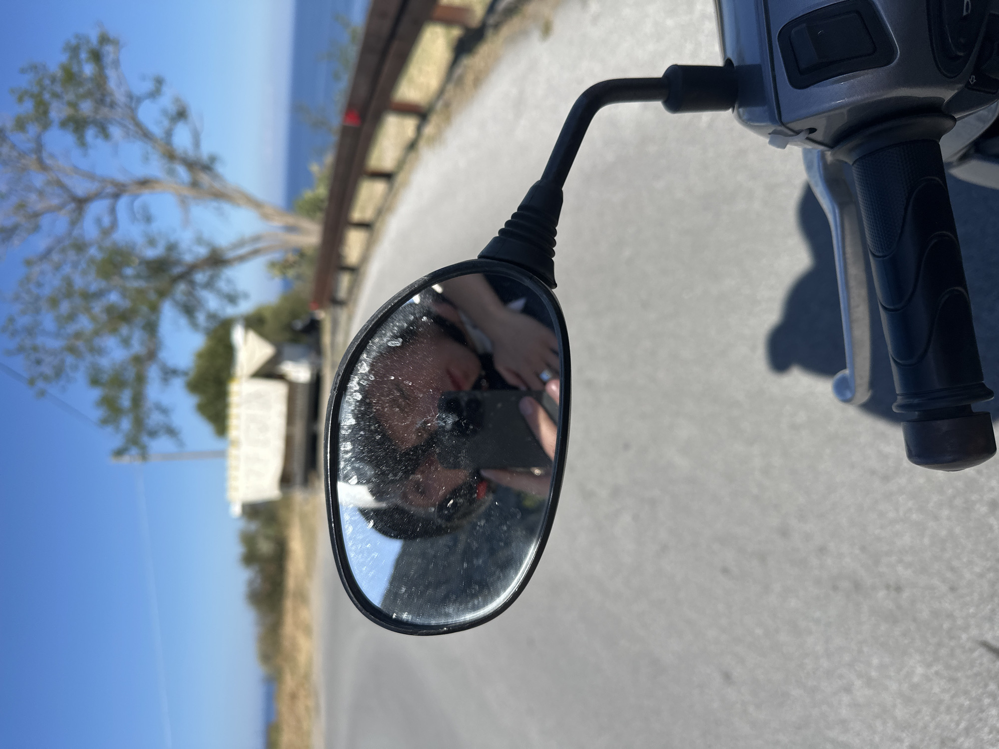 The image, rotated 90 degrees counter-clockwise, presents a first-person perspective from a scooter, centered around its rearview mirror. The mirror, slightly dirty or worn, reflects the face of the person taking the photo, who is holding a smartphone to capture the scene.
Surrounding the mirror, parts of the scooter are visible: the black stem and base of the mirror, the black handlebar grip, and a portion of the grey dashboard with some indistinct controls. A prominent shadow of the scooter falls onto the light grey road surface below.
The background features a clear, vibrant blue sky filling a significant portion of the frame. A large tree with distinct, somewhat gnarled branches and green leaves stands against the sky. In the far distance, a calm blue sea meets the horizon. Along the edge of the road, a wooden fence or railing can be seen, alongside dry, sparse vegetation and a white structure, possibly a building or tent, nestled among greenery. The road itself appears to be paved and curves gently.
The bright, clear light indicates it is daytime with sunny weather, aligning with a scenic coastal or island setting like Lipari, Italy. No discernible text is visible in the image. The overall scene captures a moment of travel or exploration, with the photographer using the mirror for a unique self-portrait.