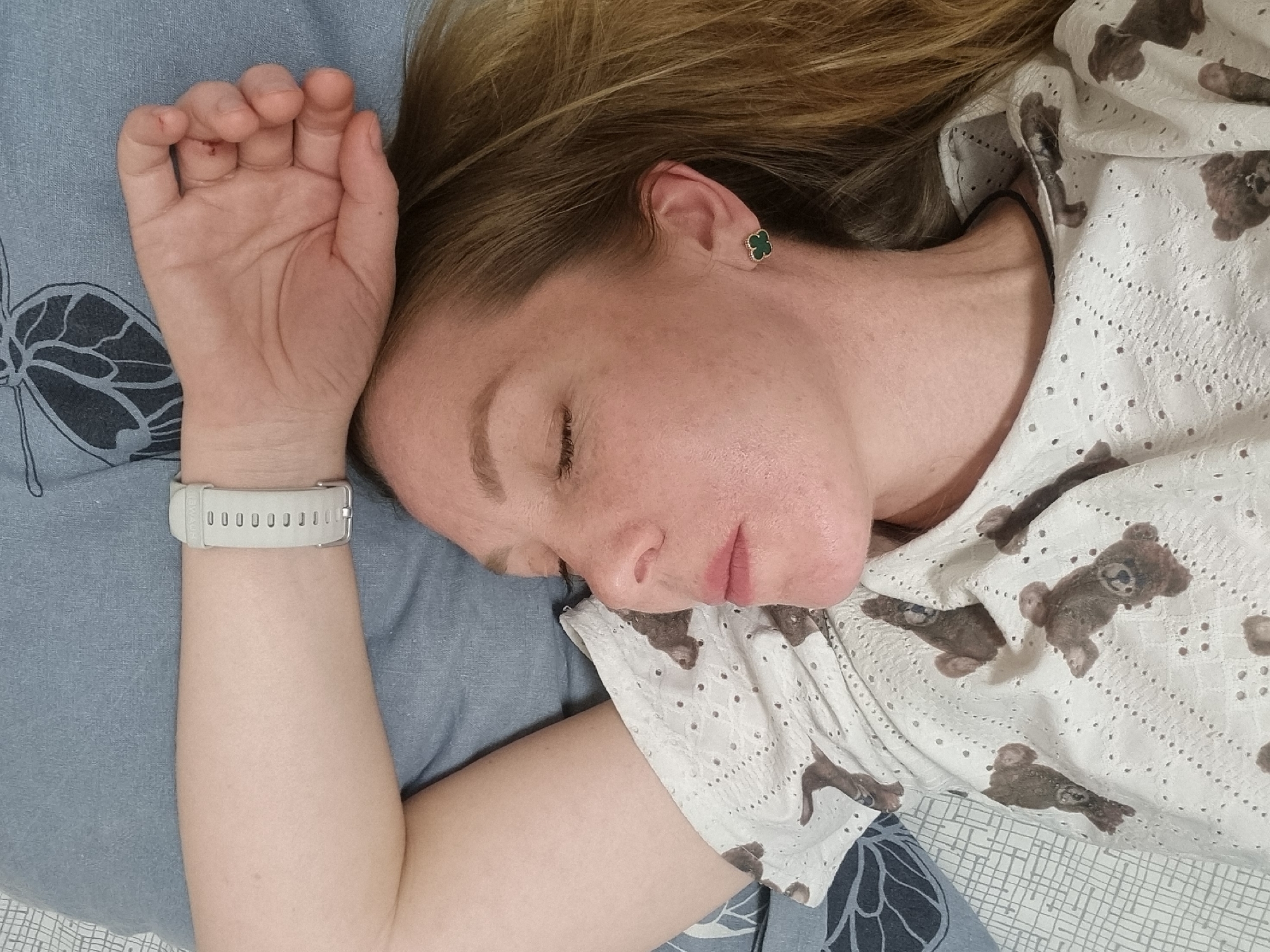 A woman is sleeping on a bed. She is lying on her side, with her head on a blue pillow. She is wearing a white top with brown teddy bear patterns. She has a white smartwatch on her wrist. She is wearing a green clover-shaped earring. The bedding is a combination of blue and white patterned fabric. There is no information available in the image to confirm the location in the city of Chaikovsky, Russia.