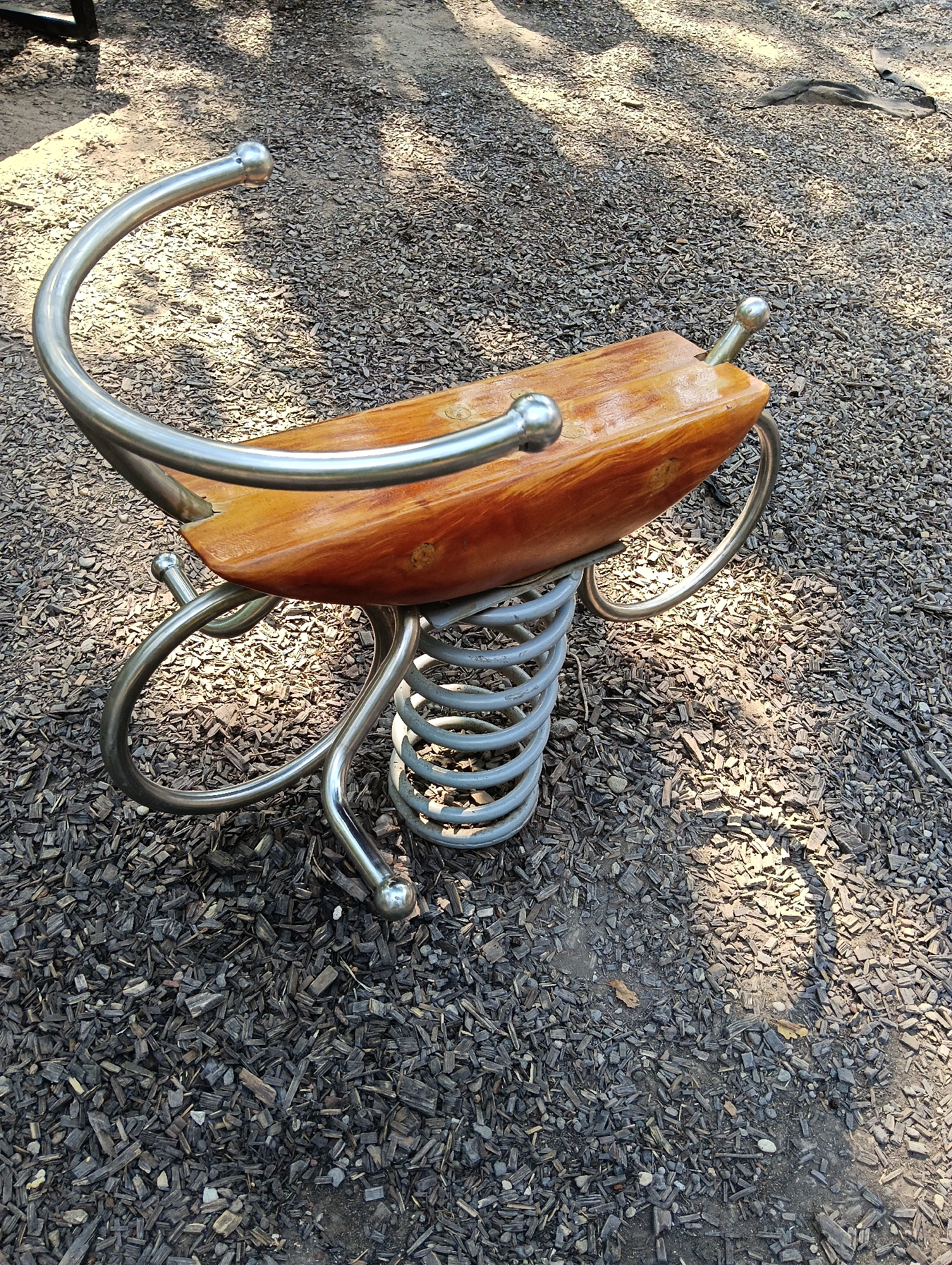 A close-up, eye-level shot depicts a wooden and metal spring rider on a playground. The rider, shaped like an abstract insect or sea creature, has a polished wooden seat with rounded edges and two chrome handlebars that curve upwards and outwards, each capped with a small spherical knob. The rider is supported by a large, grey, coiled spring attached to a circular metal base. The spring is partially obscured by the wooden seat and handlebars. The ground beneath is covered with a thick layer of dark wood chips, characteristic of a playground safety surface. Sunlight dapples the wood chips, creating patches of light and shadow, suggesting an outdoor setting during the day. The scene is clean and appears to be in good condition. No people are visible. The location is identified as Haskovo, Bulgaria.