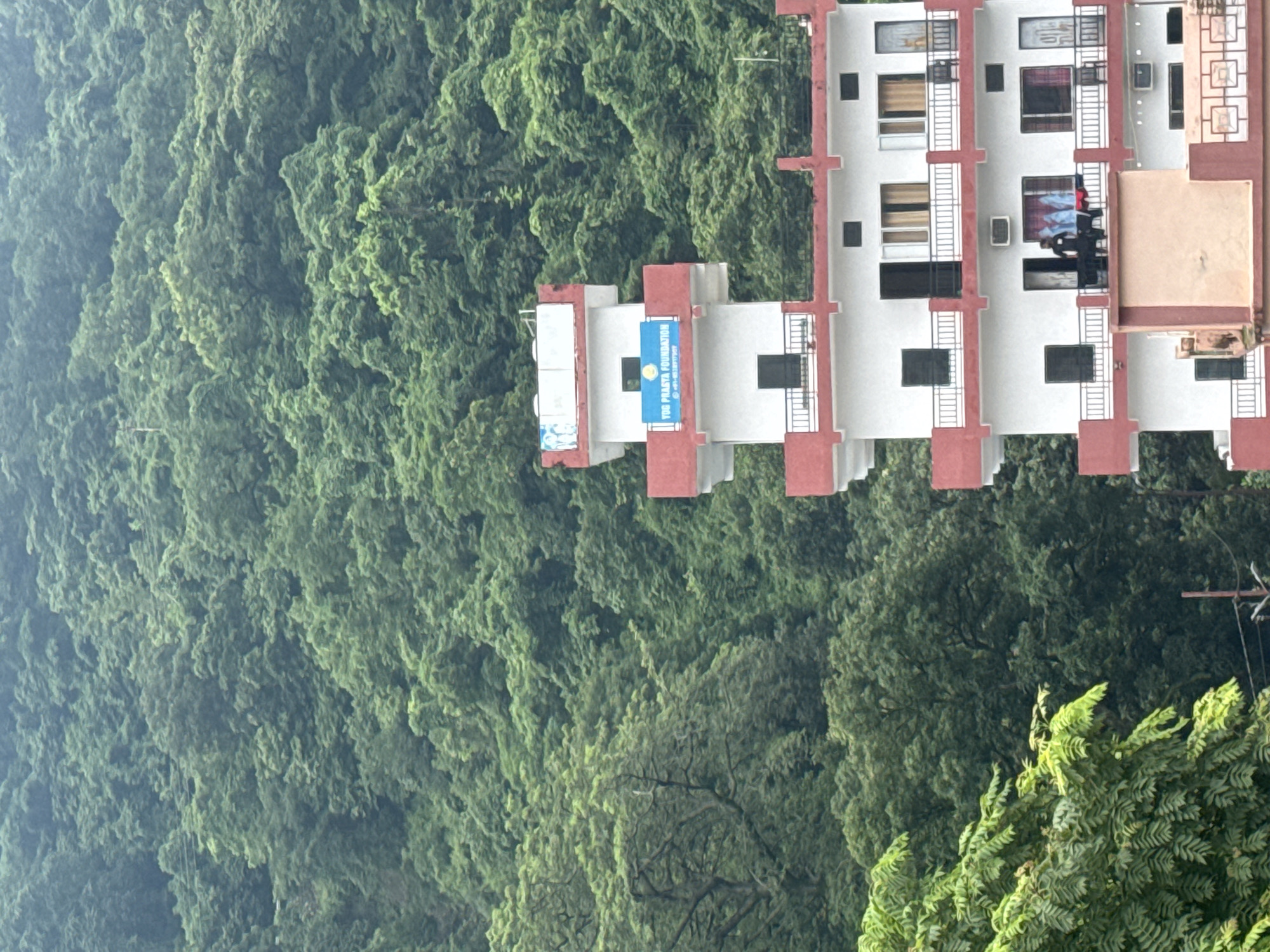 A multi-story building with white walls and red trim stands prominently in the foreground against a backdrop of lush, green trees covering a hillside. The building has multiple balconies with railings and windows. A blue sign with white text that reads "YOG PRASYA FOUNDATION" is visible on the upper section of the building. Two individuals are standing on a balcony on the third floor. The overall scene suggests a location in Rishikesh, India, given the presence of the foundation and the typical mountainous terrain.