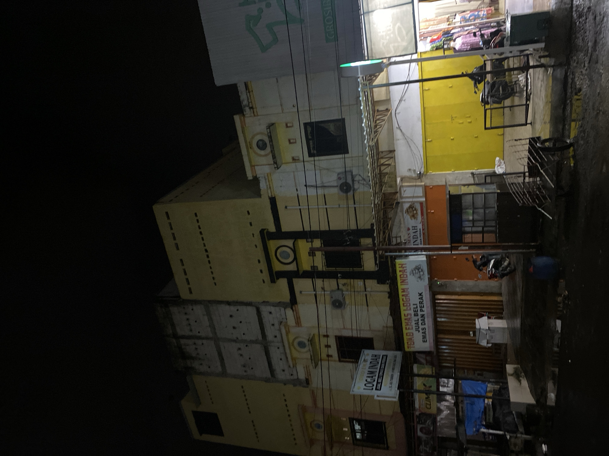 The image depicts a street scene at night in Ta', Indonesia, with a focus on commercial buildings. The main building in the center is a multi-story structure with a cream-colored facade and yellow accents. Several signs are visible, including "LOGAM INDAH" and "TOKO EMAS LOGAM INDAH," indicating a gold shop. The shop appears to sell gold and silver. In front of the shop, a rolling shutter door is closed. To the right of this building, another storefront with a bright yellow shutter is visible. Further to the right, a building with a large green logo and the word "GROSIR" (wholesale) is partially visible. The street in front of the buildings is wet, suggesting recent rain. Several motorcycles are parked in front of the shops. A small cart with metal rails is positioned on the left side of the foreground. The overall impression is of a commercial area in Indonesia at night.