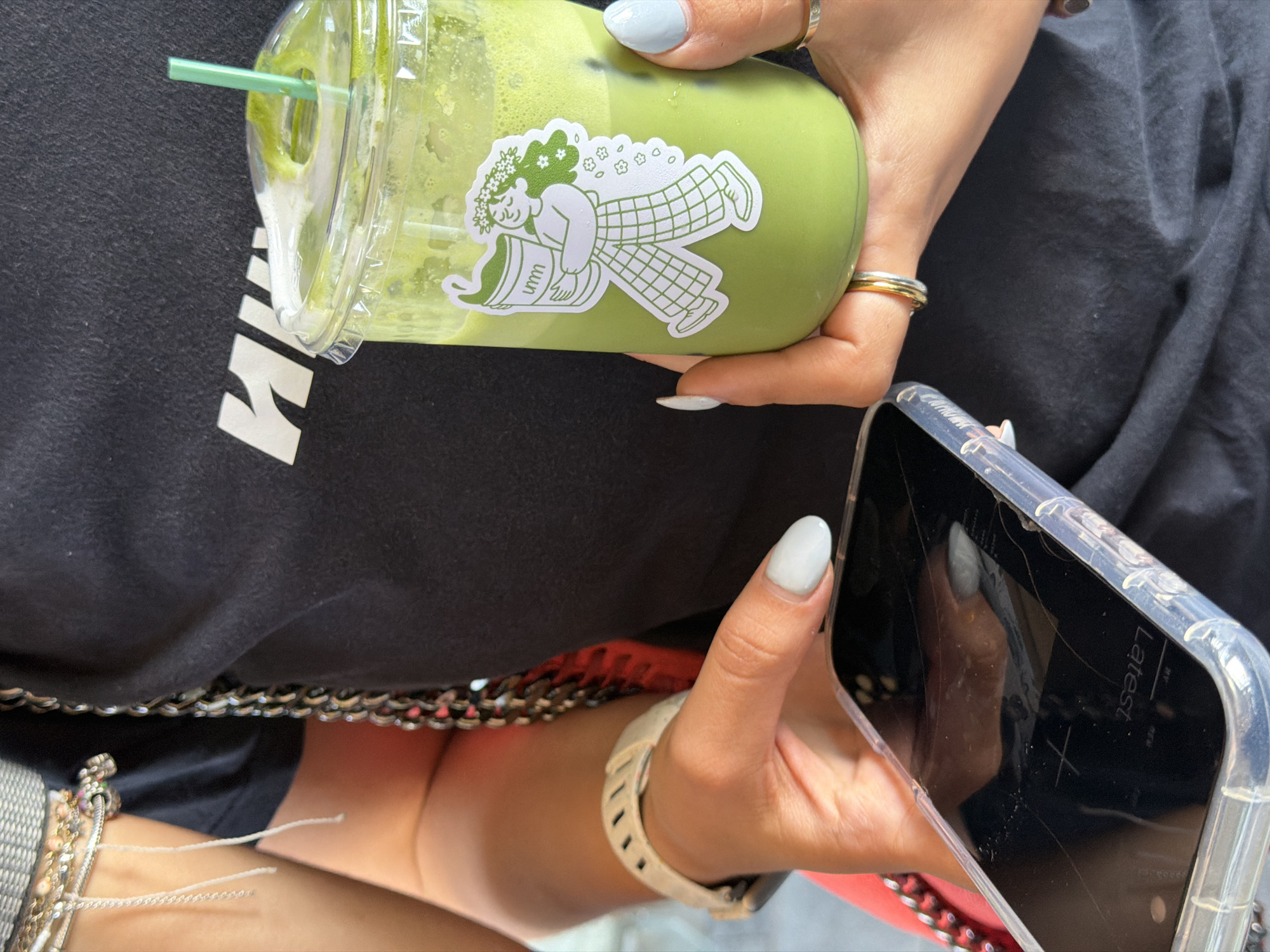 A person is holding a green drink with a sticker on it in their right hand and a cracked smartphone in their left hand. The person is wearing a black shirt with a white design on it, multiple rings on their right hand, a watch and multiple bracelets on their left wrist, and light blue nail polish. The background is blurred. The location is Puebla, Mexico.