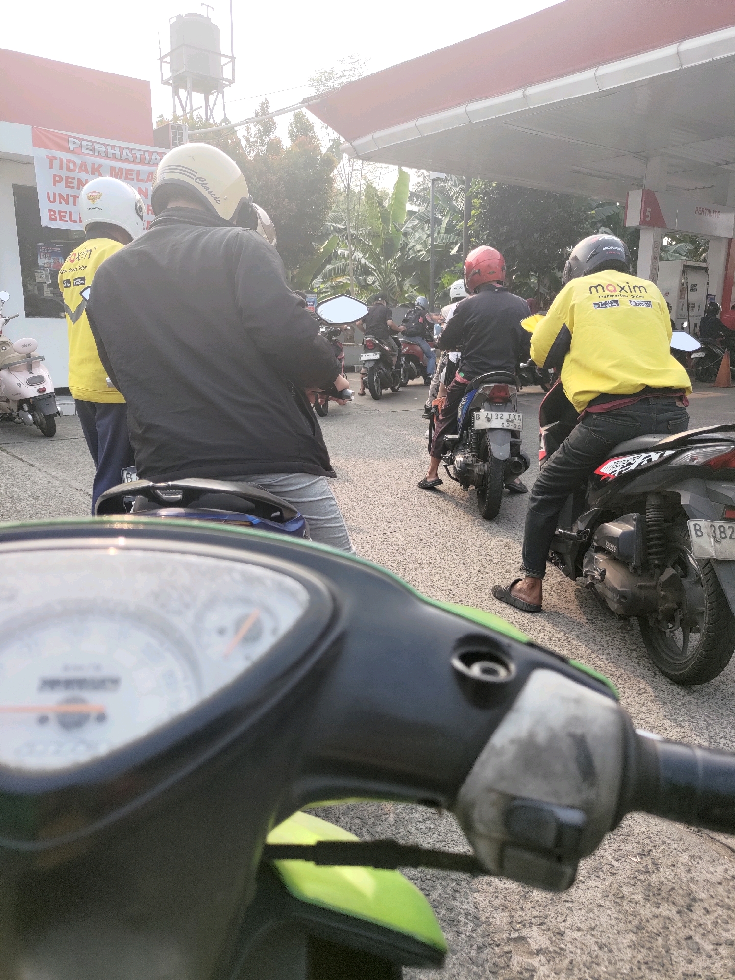 A group of people on motorcycles are queuing at a gas station in Depok, Indonesia. The scene is viewed from the perspective of someone on a motorcycle in the foreground, looking forward. Several motorcycles, some with riders, are visible in the middle ground, waiting in line. One motorcycle in the foreground is dark blue with a green front panel and a blurry instrument cluster.
In the middle ground, a person wearing a yellow shirt with "Maxim" printed on it and a black helmet is on a motorcycle. Another person in a black jacket and a yellow helmet is in front of them. To their right, a person in a red helmet and black jacket is on a motorcycle. Further back, other motorcycles and riders are visible.
The gas station canopy is visible overhead, with a sign reading "PERTALITE". A water tower and trees are in the background. The ground is paved asphalt. The overall impression is a busy scene at a gas station during daylight.