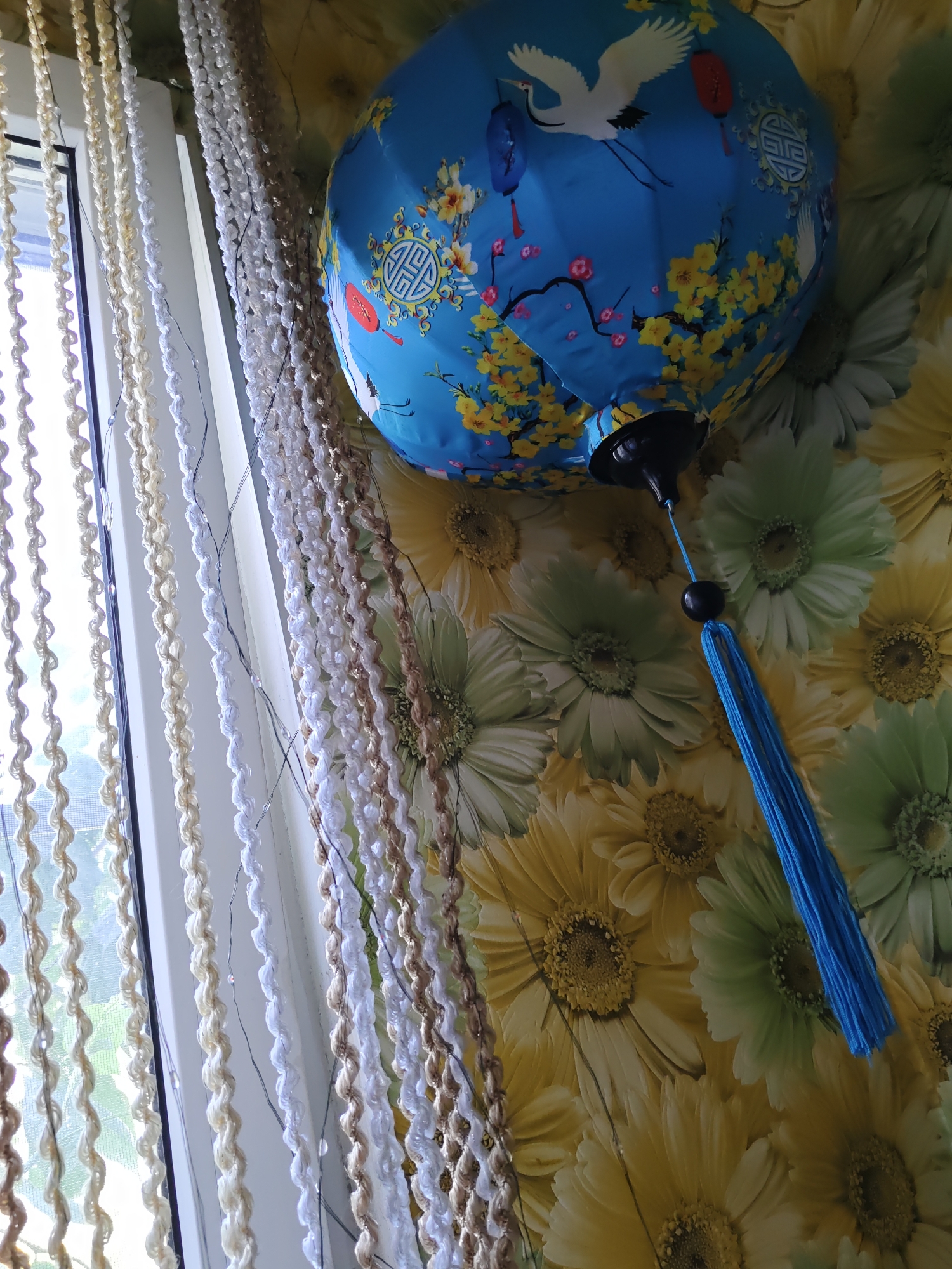 This is an indoor, eye-level, close-up shot of a blue decorative lantern hanging against a wall with a floral wallpaper pattern. The lantern is spherical with a decorative pattern of cranes, blooming branches with yellow flowers and pink blossoms, and stylized circular emblems. A black socket is visible at the bottom of the lantern, from which a black cord extends to a blue tassel. Multiple strands of decorative beaded curtains, in white, beige, and brown tones, hang in the foreground on the left side of the image. The background wallpaper features a repeating pattern of large, daisy-like flowers in yellow and green hues. The lighting suggests it is daytime, with natural light coming from a window to the left, indicated by the window frame and diffused light on the curtains. There are no people visible in the image. The scene is consistent with an interior decoration. The city of Rostov-on-Don, Russia is not depicted in this image.