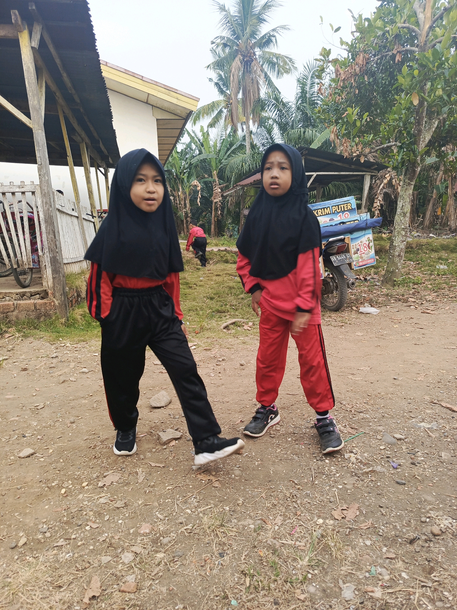 In Megang Sakti II, Indonesia, two young girls, dressed in matching red and black athletic wear and black hijabs, stand on a dirt path. The girl on the left, closer to the camera, has her hands in her pockets and is looking slightly to her left. The girl on the right is looking towards the camera with a neutral expression. Behind them, a motorcycle with a blue ice cream cart attached is parked. The cart has text that appears to read "KRIM PUTER" and a numerical "10". Further in the background, a simple wooden structure with a corrugated metal roof stands, adjacent to a weathered fence. Palm trees and other tropical vegetation are visible, suggesting a warm climate. The ground is covered in dirt, small rocks, and patches of dry grass. Another individual, wearing a red shirt and black pants, is partially visible in the background, walking away from the camera.