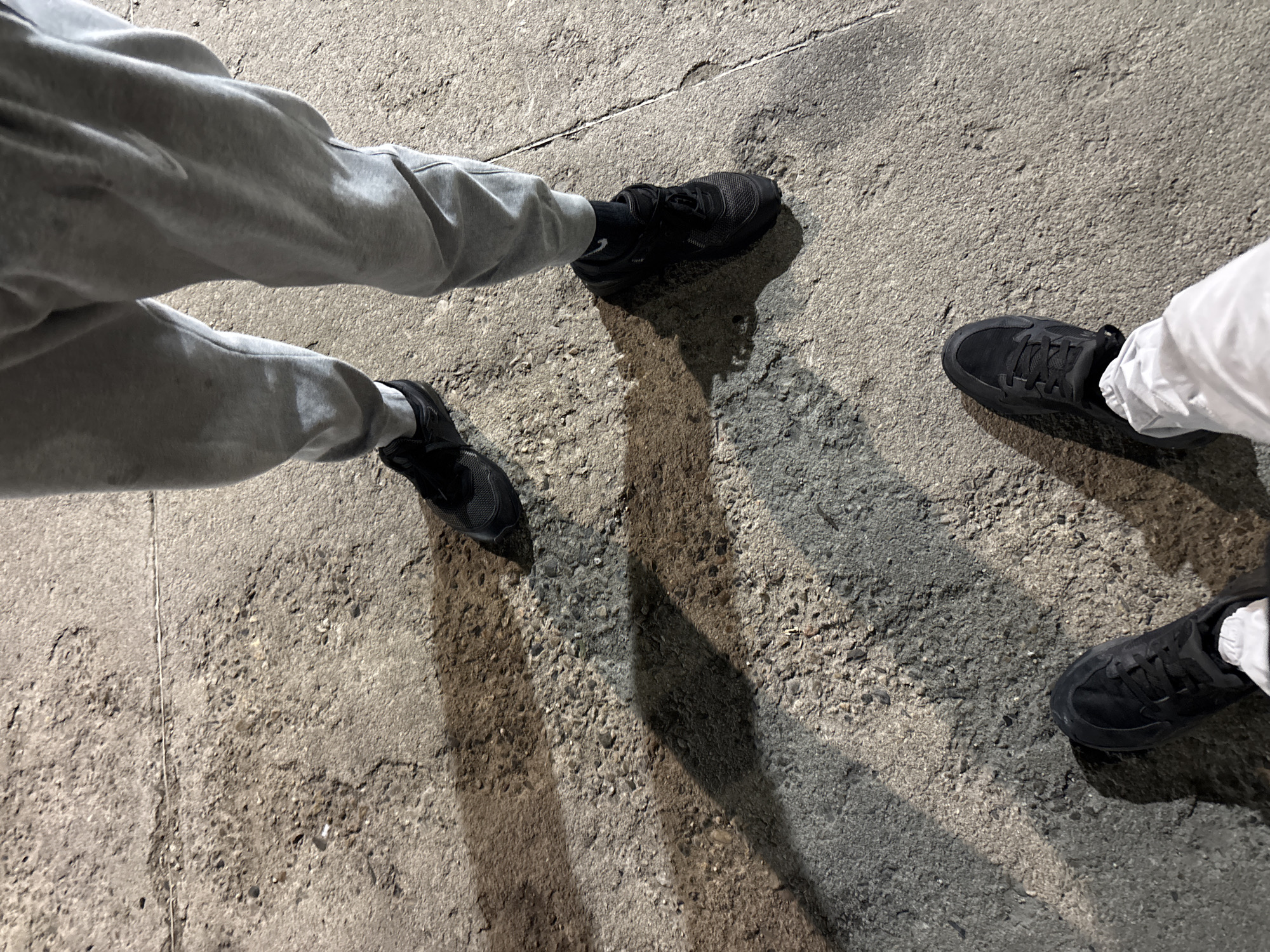 The image shows a ground-level view of two pairs of feet standing on a concrete surface. The person on the left is wearing grey sweatpants and black sneakers with dark socks. The person on the right is wearing white pants and black sneakers with white socks. The concrete surface is textured and cracked. The lighting is dim, casting shadows from the feet and legs. The location cannot be confirmed. There are no other visible objects.