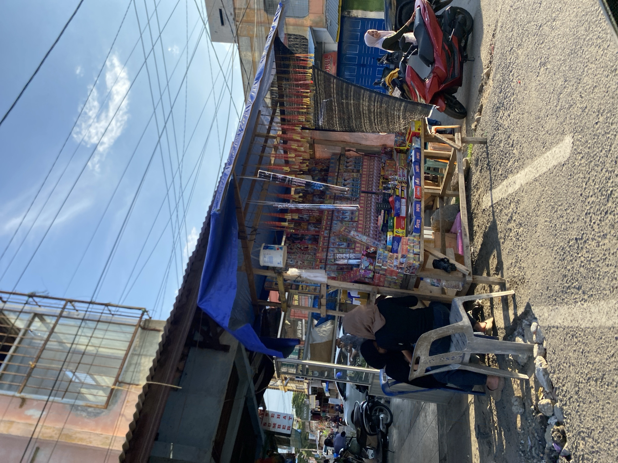 In Lhokseumawe, Indonesia, a roadside stall displays fireworks, firecrackers, and various packaged goods for sale. The stall is constructed of wooden frames and is partially covered with a blue tarp. A woman wearing a hijab sits in a plastic chair nearby. A red motorbike is parked next to the stall on the paved road. Overhead, power lines crisscross against a blue sky with sparse clouds. Other buildings and businesses are visible in the background along the street.