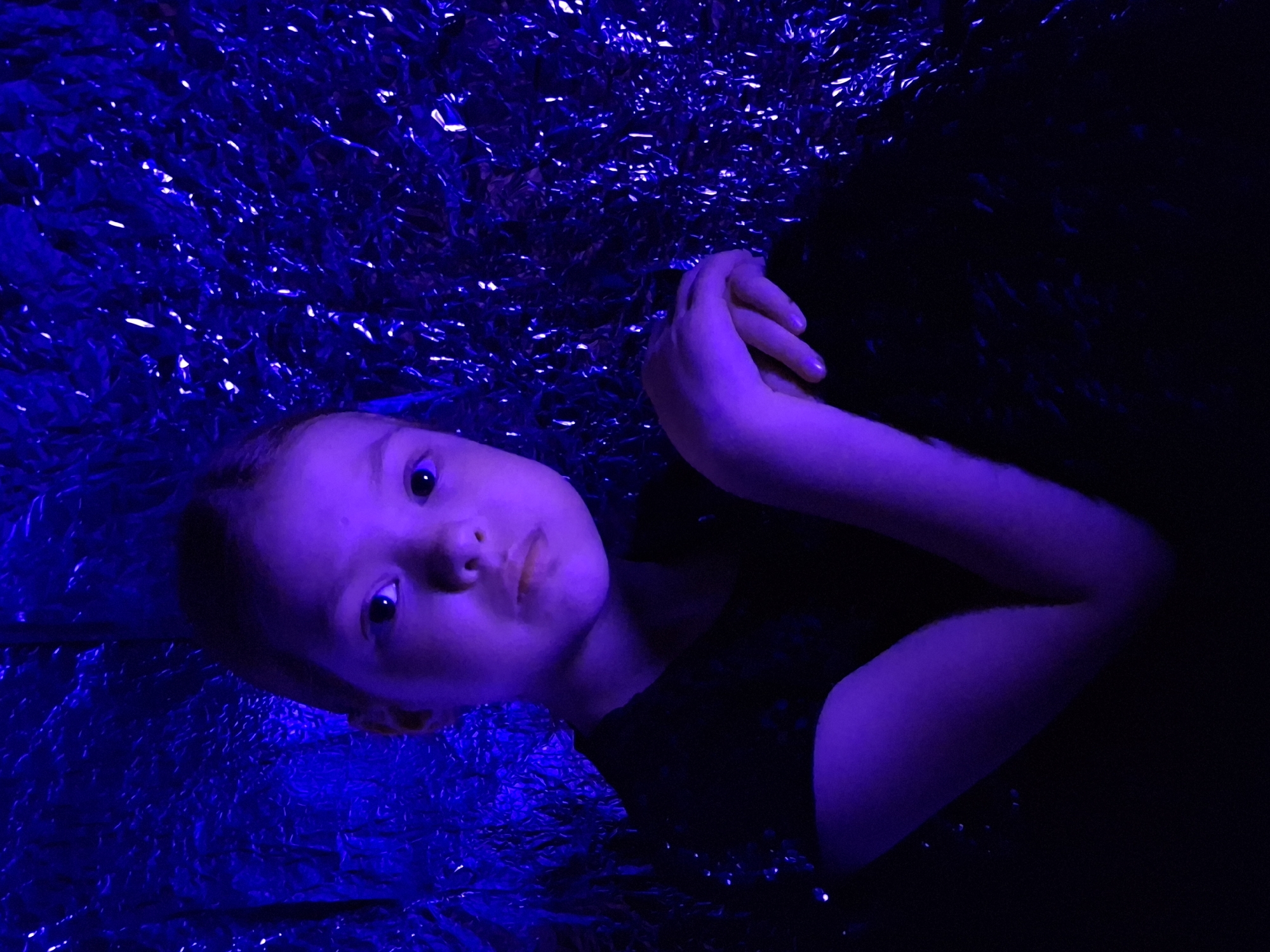 A child lies on a surface of crumpled foil or similar material. The scene is lit with a strong, blue light that casts deep shadows. The child is wearing a dark garment. Their hands are folded near their face. The location is unknown but the prompt suggests it is in the city of Chaikovsky, Russia.