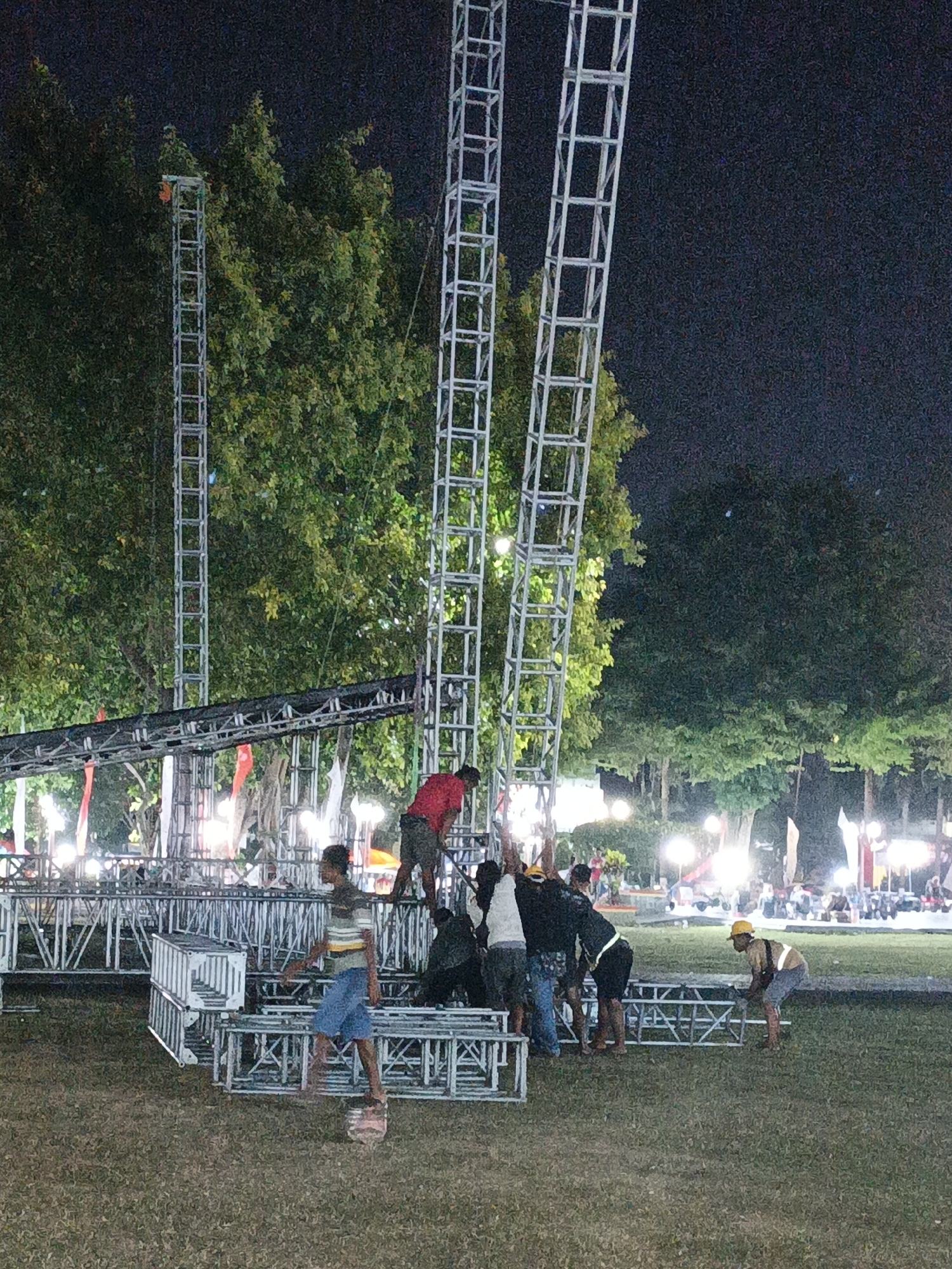 In Klaten, Indonesia, at night, several individuals are actively engaged in assembling a large metal truss structure on a grassy outdoor field. Three tall, vertical truss towers are visible, along with horizontal truss components and what appear to be stacked modular truss sections. A group of men are clustered around the base of the towers, some appearing to be lifting or connecting pieces. One man, wearing a red shirt and dark shorts, is bent over near the structure. Another individual, in a striped shirt and blue shorts, is walking away from some of the assembled truss elements. In the background, numerous bright lights illuminate the scene, suggesting a public event or performance is being prepared. Trees with dense green foliage form a backdrop against the dark night sky. Further in the distance, a crowd of people and additional lighting indicate a larger gathering is present. One person on the right side of the frame is wearing a yellow hard hat and appears to be working with the truss.