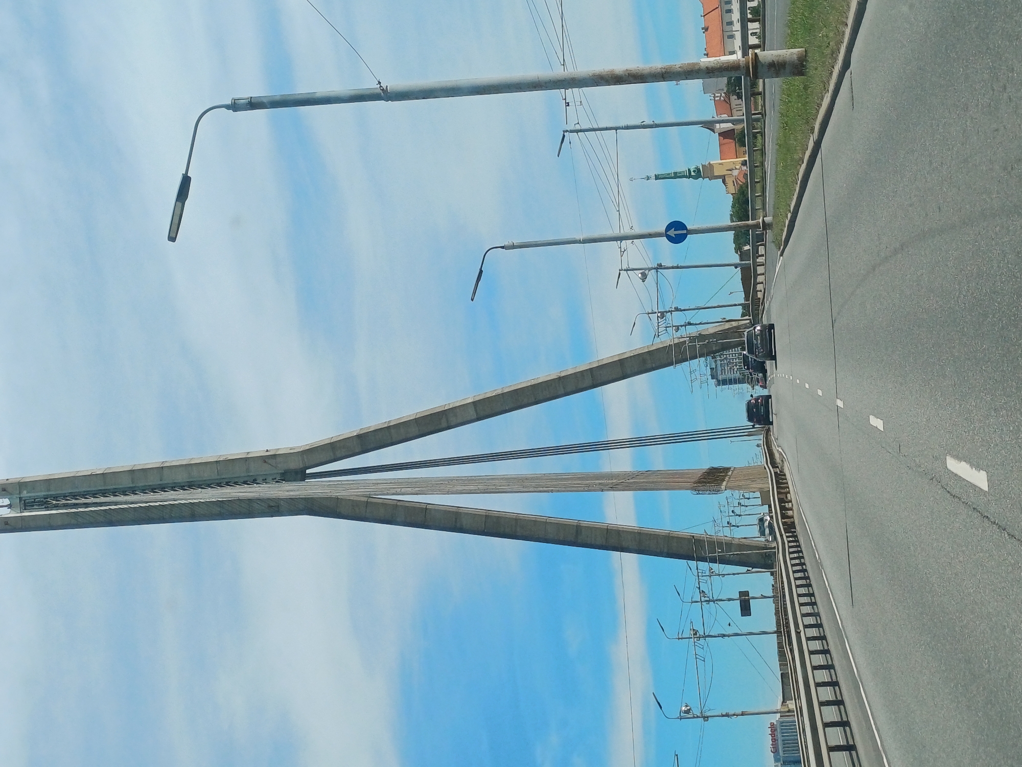 The image shows a view from a vehicle driving on the Vanšu Bridge in Riga, Latvia. The bridge's distinctive tall concrete pylons and cable supports are visible against a blue sky with scattered clouds. The road ahead is asphalt with white lane markings, and guardrails line the sides. Several vehicles, including cars and a truck, are on the bridge. In the background, buildings with orange roofs and a church spire are visible on the city's skyline. Streetlights are mounted on poles along the bridge. The scene depicts a sunny day on a major city bridge.