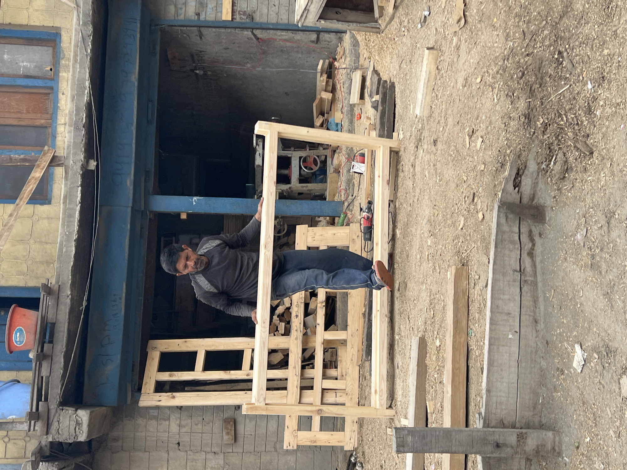 A man is standing in a cluttered outdoor workshop in Dras, India, holding a large wooden frame. He is wearing a dark grey long-sleeved shirt and blue jeans, with orange-soled sandals. The frame appears to be part of a bed or a similar structure. Wood scraps and tools, including a red and black power drill, are scattered on the ground around him. In the background, a workshop structure is visible with more wooden materials and machinery. The scene suggests ongoing construction or woodworking activity in a somewhat unorganized environment.