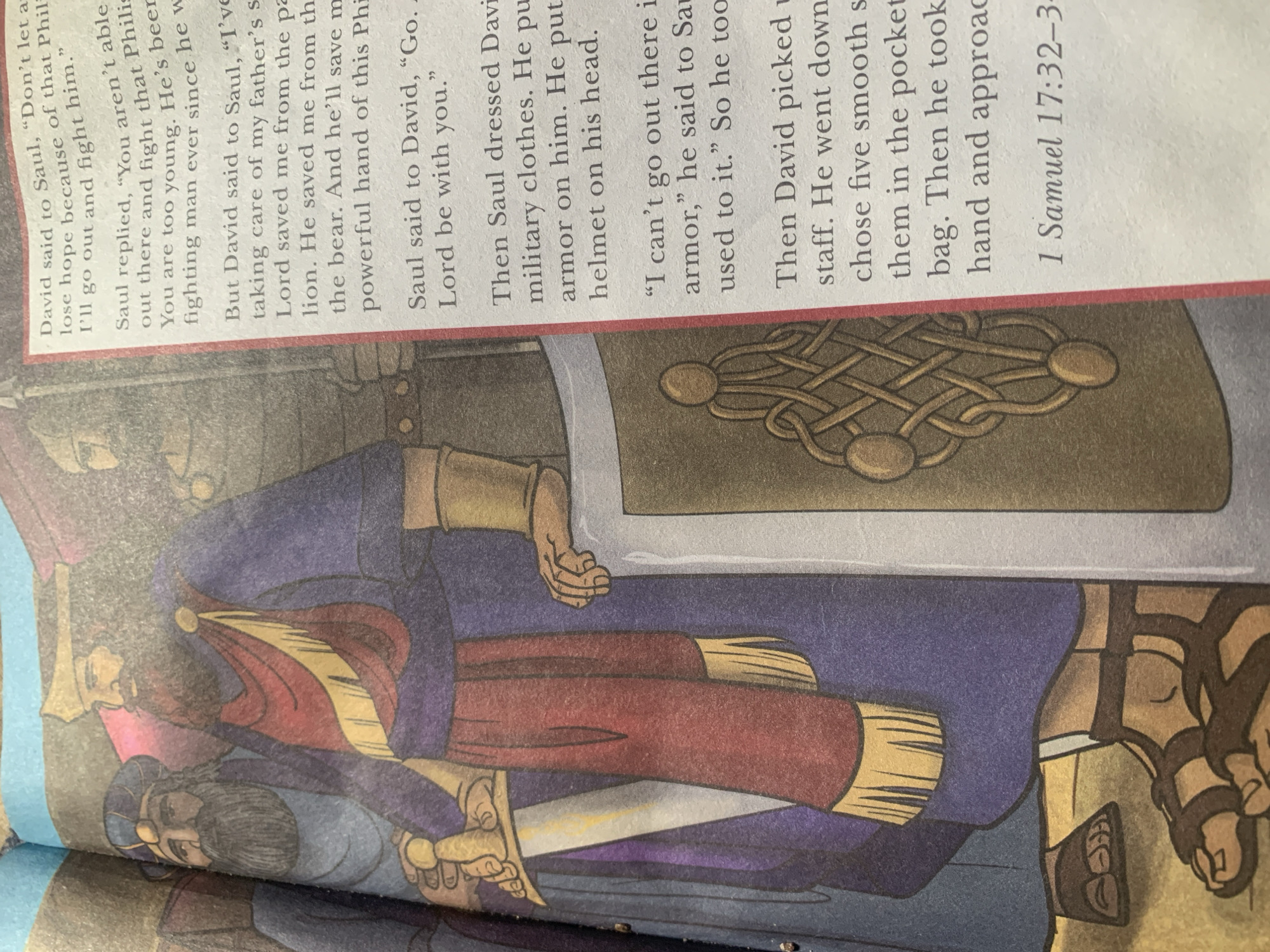 The image shows an illustration from a book, depicting a scene with two men in historical attire. One man, presumed to be David, stands to the left, holding a sword. He wears a blue tunic and a red sash with gold trim. Another man, likely Saul, stands beside him, wearing a purple robe and a gold crown. The background is a muted, textured wall. The text on the right side of the page is from the Bible, specifically 1 Samuel 17:32-34, detailing a conversation between David and Saul. There is no indication of the city of Jauro Yinu, Nigeria in the image.
