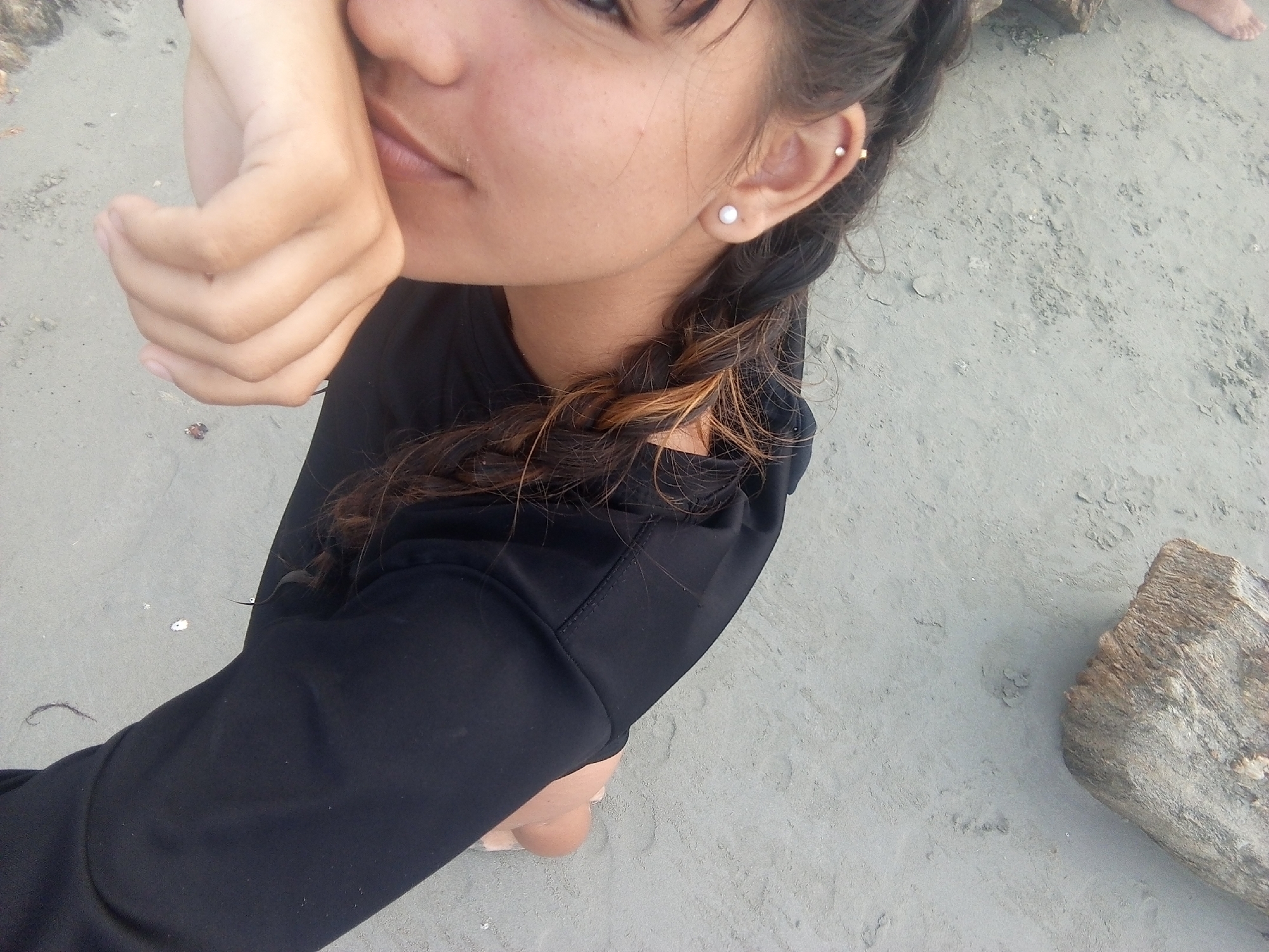 A woman is pictured outdoors on a sandy surface, possibly a beach in the city of Juan Griego, Venezuela. She is wearing a long-sleeved black garment and has braided hair with some blonde highlights visible. She has two earrings on her left ear. She partially covers her face with her hand. The sandy ground is light gray and there is a large piece of driftwood nearby on the right side of the frame. Part of a bare foot can be seen behind the woman.