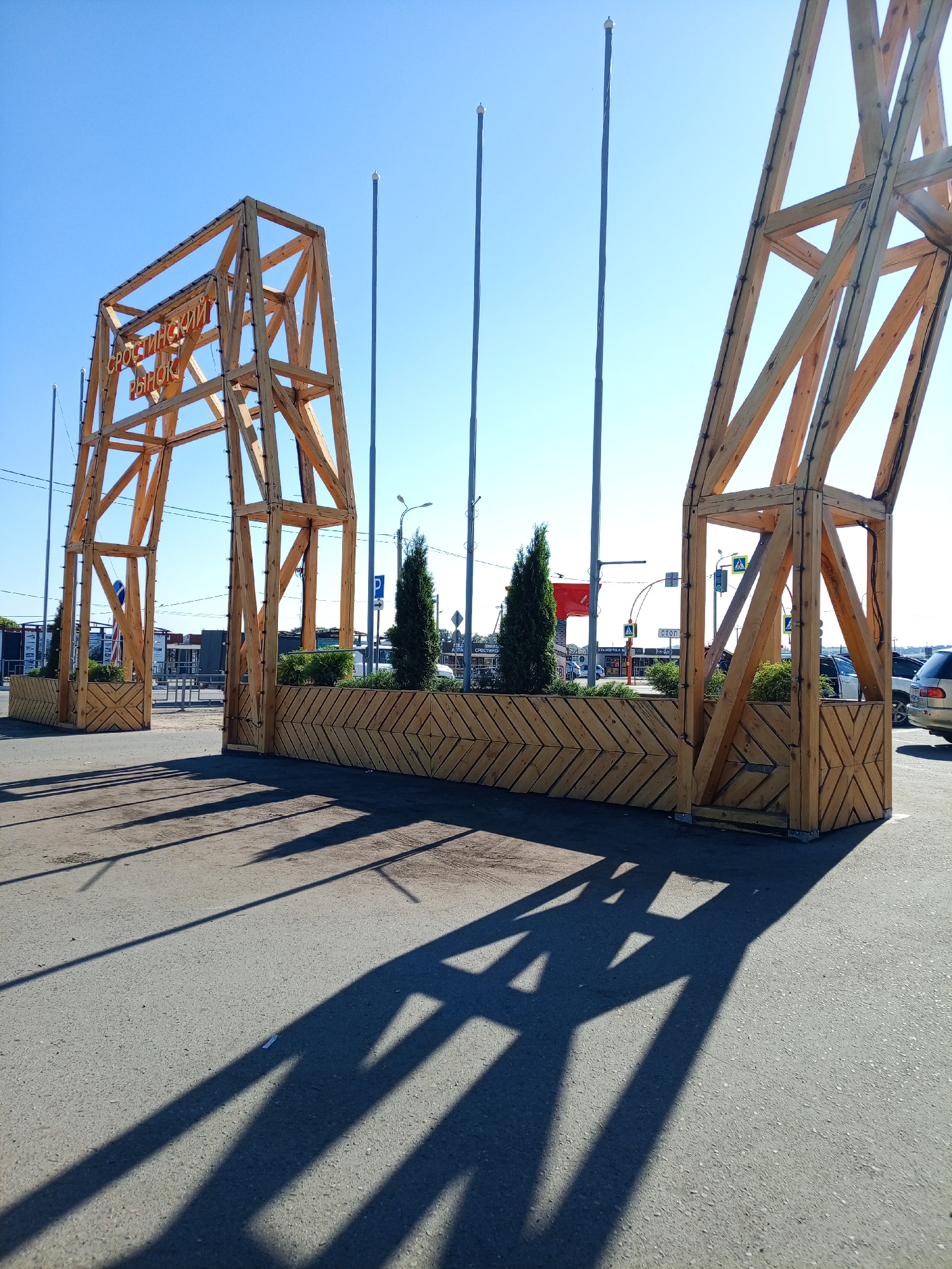 The image shows a large wooden archway, likely an entrance to a market or event, in Srostki, Russia. The arch is constructed of light-colored wood beams forming a geometric structure. Red lettering, partially obscured by the wooden frame, is visible on the top part of the archway, reading "СРОСТИНСКИЙ РЫНОК" (Srostinskiy Rynok), which translates to "Srostki Market." The archway is situated outdoors on a paved surface, possibly a road or parking lot, under a clear blue sky. The sun is casting long shadows from the wooden structure onto the ground, indicating it is likely midday or afternoon. In the background, several tall, thin poles or flagpoles are visible. Behind the archway, there are some low green bushes planted in wooden planters. Further in the background, traffic signals, streetlights, and buildings that appear to be part of a commercial area can be seen. A few vehicles are also present in the background. The overall scene suggests a sunny day in a public outdoor space in Srostki.