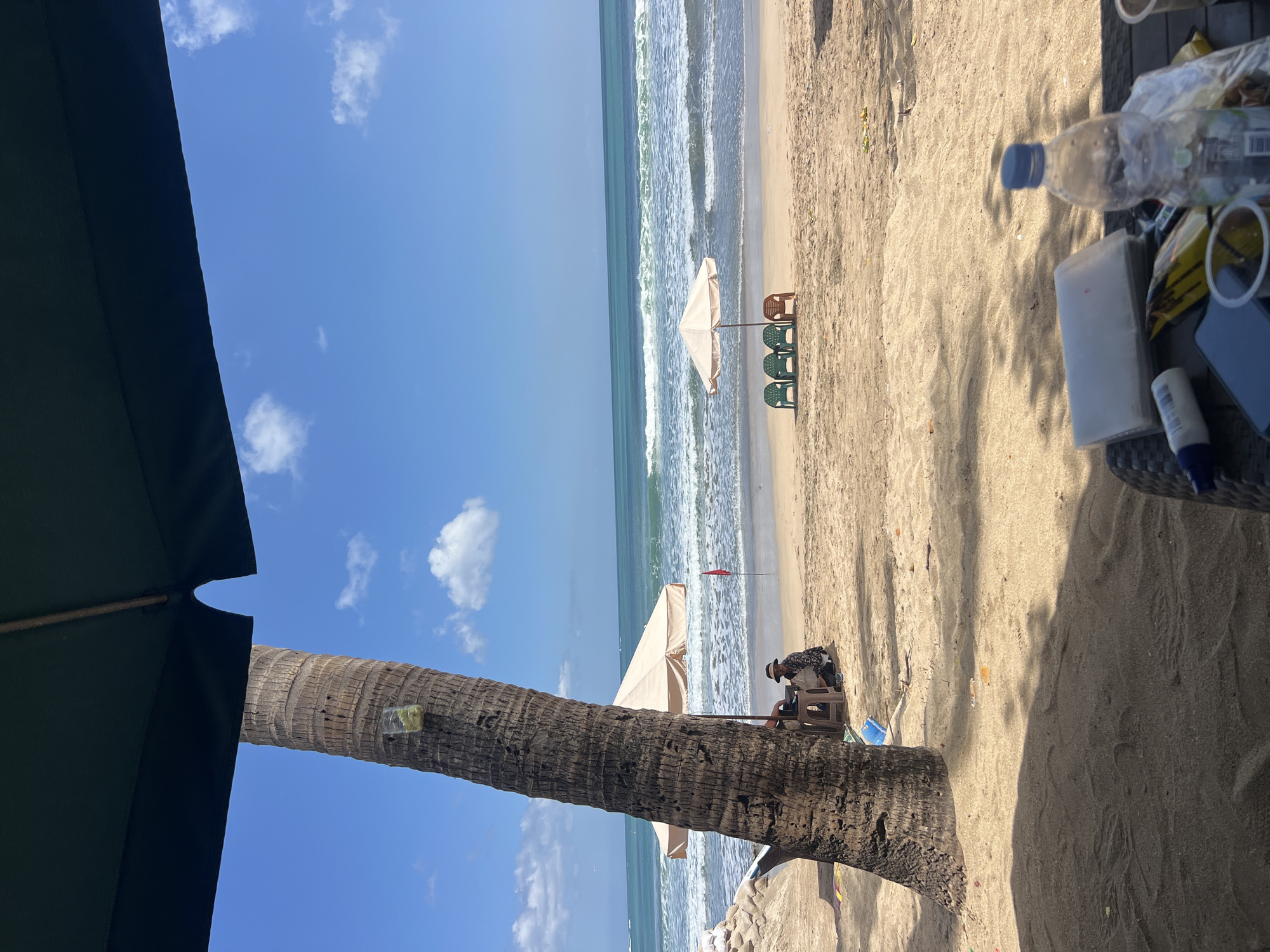 The image depicts a sunny day at Kuta Beach, Indonesia. In the foreground, a portion of a dark green umbrella shades a wicker table with various items, including a plastic water bottle, a phone, and a stack of papers. To the left, a tall palm tree trunk with a clear plastic cup attached to it stands prominently. Further back, a person wearing a hat and a patterned shirt sits on a brown stool next to a light-colored beach umbrella. Beyond them, the sandy beach stretches to the ocean, where gentle waves roll onto the shore under a clear blue sky with scattered white clouds. A line of three green plastic chairs and one brown plastic chair are visible near another beach umbrella, and a red flag is planted in the sand further down the beach. The overall scene is a serene beach setting in Kuta, Indonesia.