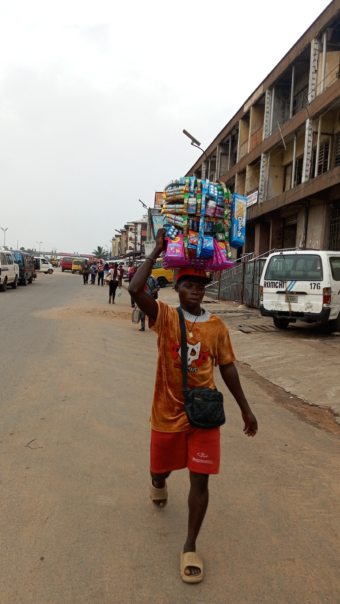 The image depicts a vibrant street scene in Onitsha, Nigeria, under an overcast sky, suggesting daytime. The central subject is a young male street vendor, walking towards the viewer. He expertly balances a large, rectangular tray piled high with numerous packaged snacks, crisps, and candies on his head. He wears an orange t-shirt featuring a stylized graphic and the word "WOLFE," red shorts with "Aspresent" printed on the left leg, and light-colored slide sandals. A black shoulder bag is worn across his chest. His expression is neutral and focused.
The setting is a busy urban street with an asphalt surface. In the background, numerous vehicles, including vans and mini-buses typical of the region, are parked or moving along the road. Several pedestrians are also visible walking in the distance. On the right side of the street, a multi-story building with a weathered facade and exposed balconies lines the road, with other commercial or residential structures extending further. A white van parked on the right prominently displays the text "ROMCHI," the number "176," and "NK 5897G" on its rear. Partially visible signs on buildings include "TRANSIT LTD," "FLAVORS," and fragments like "YAN..." and "YOF...". The scene portrays the daily activity and commerce characteristic of an urban Nigerian environment.