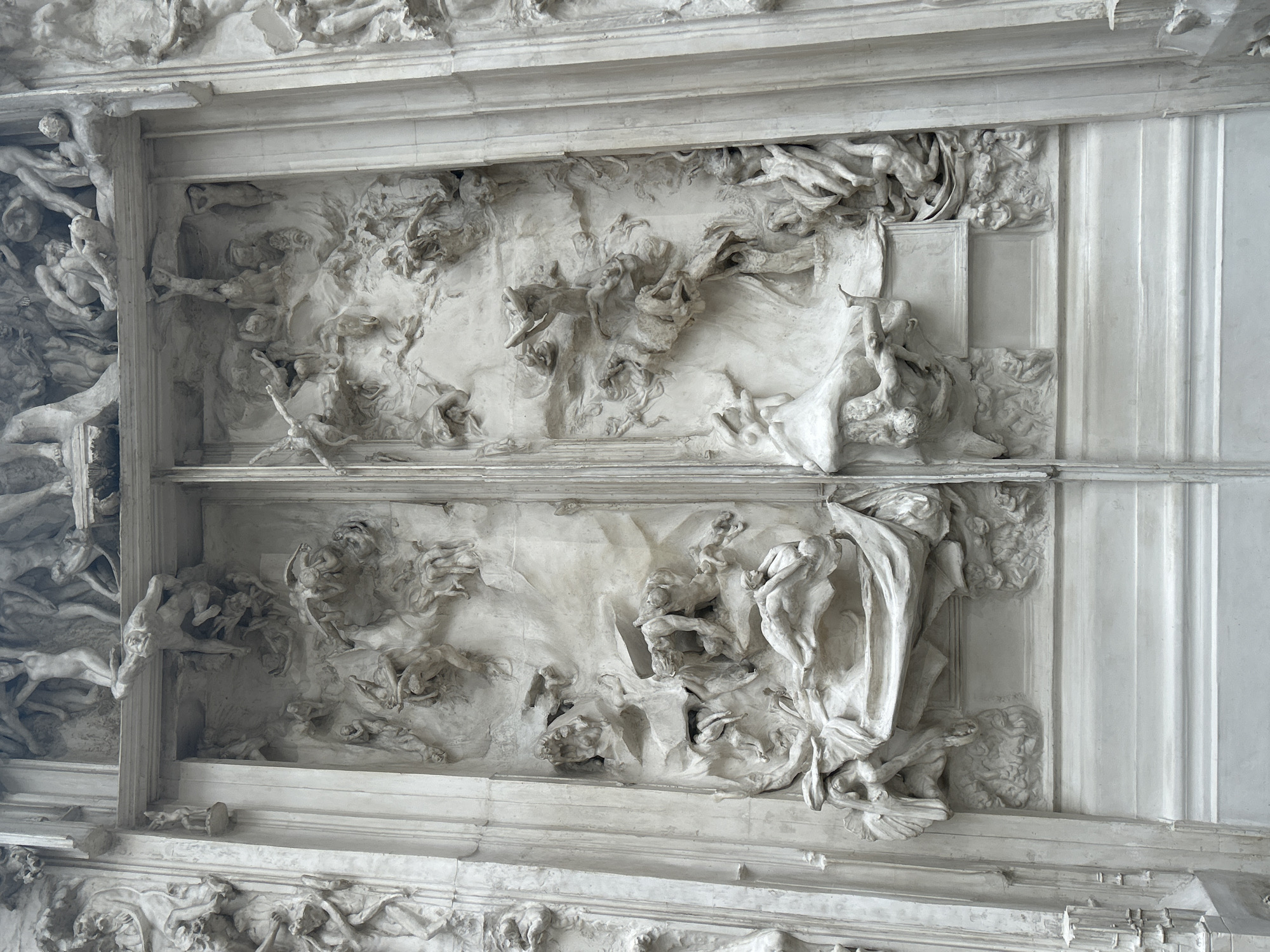 The image shows an elaborate, ornate plaster relief sculpture, likely part of a ceiling. The sculpture contains figures in dynamic poses, some draped in fabric, in what appears to be a chaotic or dramatic scene. The relief is compartmentalized into two main panels and surrounded by a decorative frame. The background is a pale, off-white color. The sculpture is located in Paris, France, specifically at the Panthéon.