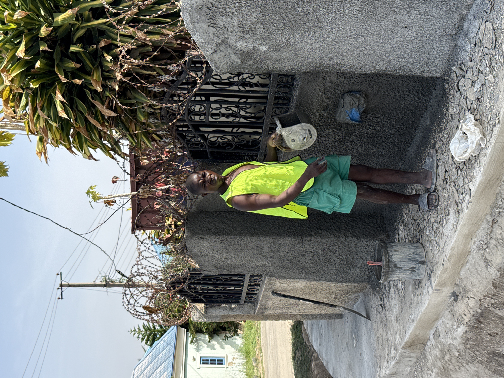 An outdoor scene captured in bright daylight shows a dark-skinned man standing in what appears to be a construction or renovation area. The man, positioned slightly to the right of the frame, wears a bright, neon yellow or lime green sleeveless vest and green shorts. He holds a small, light-colored bucket or container in his right hand and looks directly at the camera with a neutral expression.
Behind the man is a grey, textured concrete wall or pillar, partially revealing an ornate black wrought iron gate. Coils of barbed or razor wire are prominently displayed along the top of the wall and gate, suggesting a focus on security. To the left, a large, lush green plant with variegated leaves cascades downwards, partially obscuring the background.
The ground around the man features fresh grey concrete or cement work, with patches of gravel and loose debris. A dark bucket and a shovel or spade are visible on the ground near his feet. In the background, under a clear blue sky, a wooden power pole with utility lines extends upwards. Other distant structures and sparse vegetation are also visible, hinting at an urban or semi-urban environment. The bright sun casts visible shadows, indicating it is likely midday or early afternoon. No legible text is visible in the image.