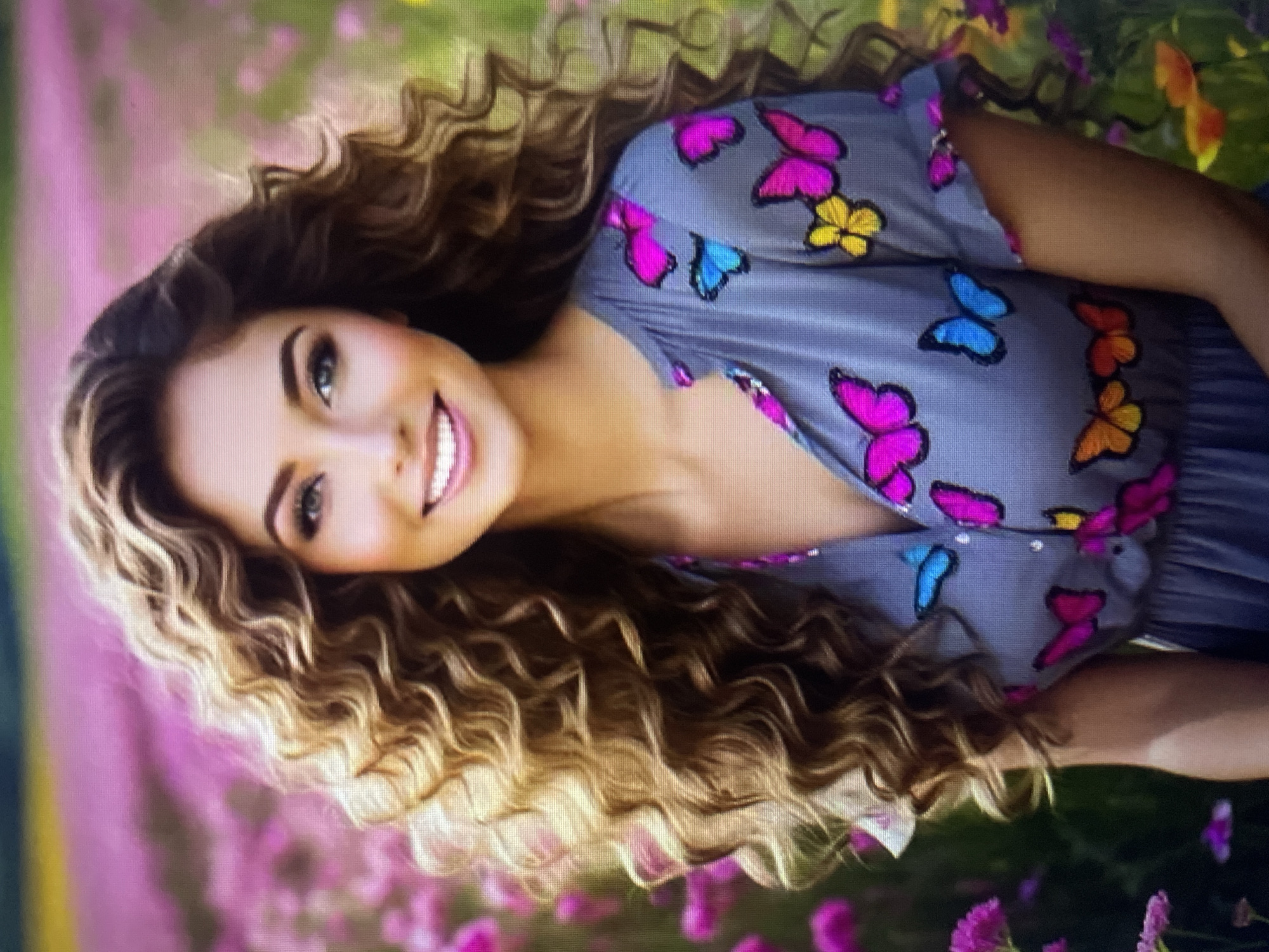 A young woman with long, curly brown hair is pictured in a field of pink flowers. She is wearing a gray dress with colorful butterfly appliques. She is smiling and looking towards the camera. The background is a blur of pink and green, suggesting a meadow or garden. There is no indication of the city of Kołobrzeg, Poland.