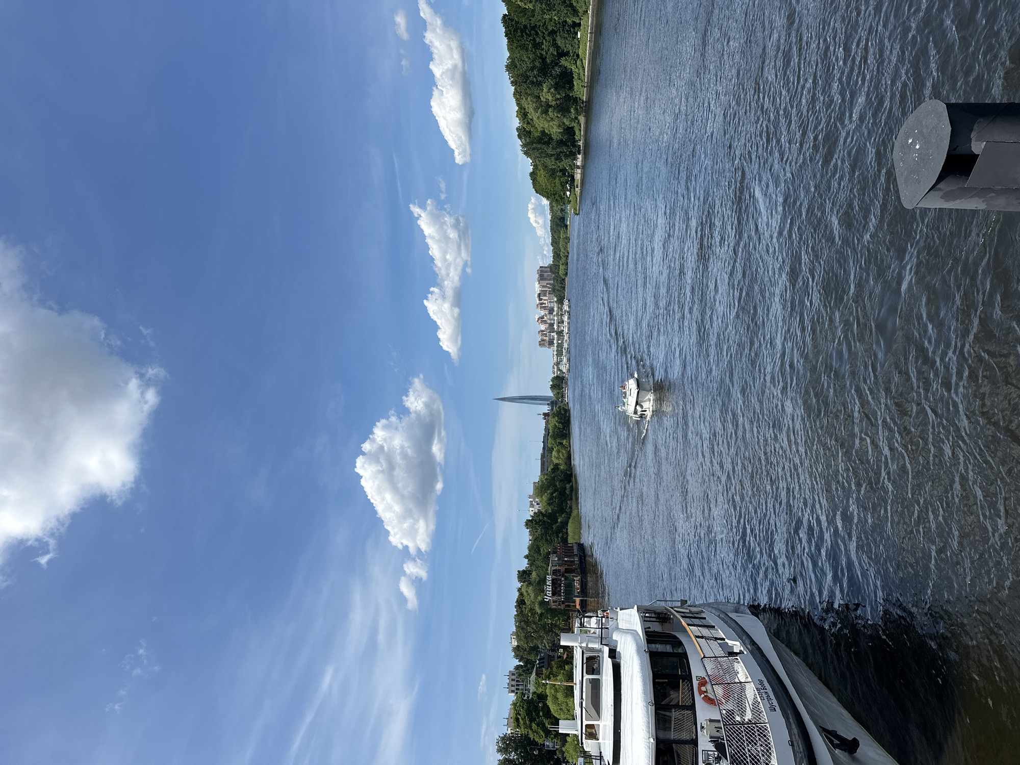 This media file captures a scenic view of a wide river in Saint Petersburg, Russia, on a bright, sunny day. The sky is a vibrant blue, dotted with fluffy white clouds. The river's surface is a deep, shimmering blue, reflecting the sky and creating a rippling texture.
The scene is dominated by water and sky, with the river occupying the right half of the frame and the sky the left. Along the far bank of the river, a lush green treeline is visible, interspersed with buildings. A tall, pointed skyscraper, the Lakhta Center, stands out against the horizon, a prominent landmark of Saint Petersburg.
Several boats are present on the water. In the foreground, a large white passenger boat is docked or moving slowly. Further down the river, a smaller white boat is seen in motion, creating a wake. Another boat with "РЕСТОРАН" (RESTAURANT) visible on its side is also present. A person in a white life vest is visible on the deck of the foreground boat.
The overall impression is one of a pleasant summer day, likely midday, with good weather. The calm water and clear sky suggest a peaceful atmosphere. The presence of boats and the cityscape indicate a populated and active area.