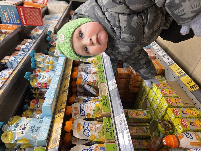 The image depicts a young child positioned in the foreground of a supermarket aisle in Chaikovsky, Russia. The child, wearing a light green knitted beanie with small protrusions and a dark gray camouflage-patterned winter jacket over light gray gloves, looks directly at the camera. Their right gloved hand is extended forward, appearing to touch or select a small green juice box from a lower shelf. The shelves behind the child are densely stocked with various baby and children's food products. The upper shelves display pouches of "ФрутоНяня" (FrutoNyanya) fruit purees, while the middle shelf holds "Агуша" (Agusha) purees in similar pouches, many labeled "100% НАТУРАЛЬНО" (100% NATURAL). The lower shelves are filled with small rectangular "Простоквашино" (Prostokvashino) juice boxes in green and orange packaging. Yellow and white price labels, displaying values in Russian rubles (e.g., 44.99, 59.99), are affixed to the front edges of the shelves. A red and white promotional sign for "yokimi.ska.ru" is visible on an upper shelf in the background. The environment is brightly lit, consistent with a retail store.