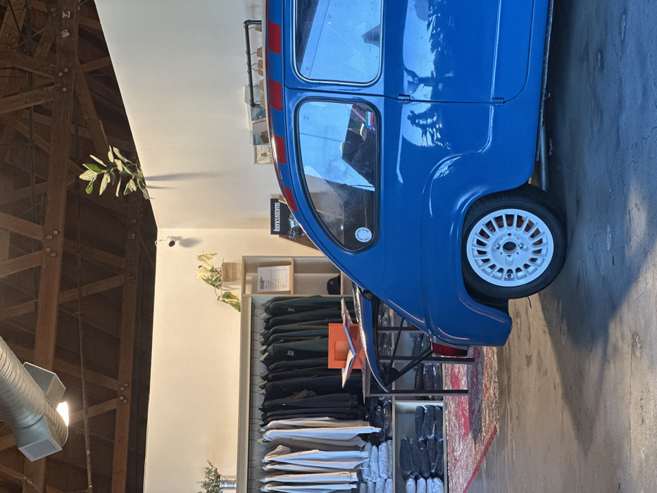 A blue car is parked indoors near clothing racks. The vehicle is on the right side of the image and close to the viewer. The clothing racks on the left showcase various garments, including shirts and pants. The ceiling is visible on the left, with an exposed wooden beam structure. A security camera is attached to the wall. A plant is visible by the wall near the ceiling.