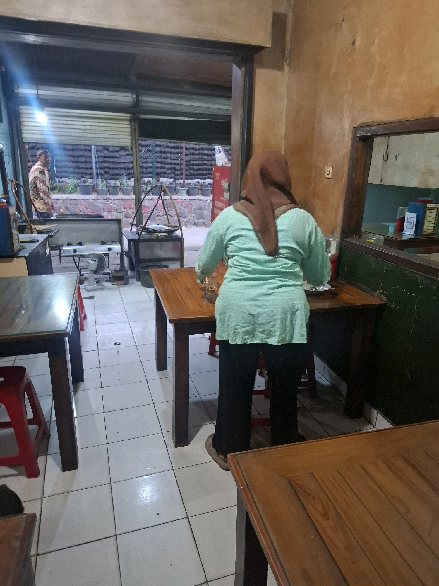 The image depicts a casual dining area within a restaurant in Citeureup, Indonesia. A woman wearing a brown hijab and a light blue tunic stands with her back to the camera, preparing food at a wooden table. In the background, through an open doorway, a man in a patterned shirt stands near a stove. The restaurant has tiled floors and wooden tables. Various kitchen appliances, including a gas stove and a fan, are visible. Shelving units are also present, holding containers and bottles. The overall scene suggests a functional, working restaurant environment.