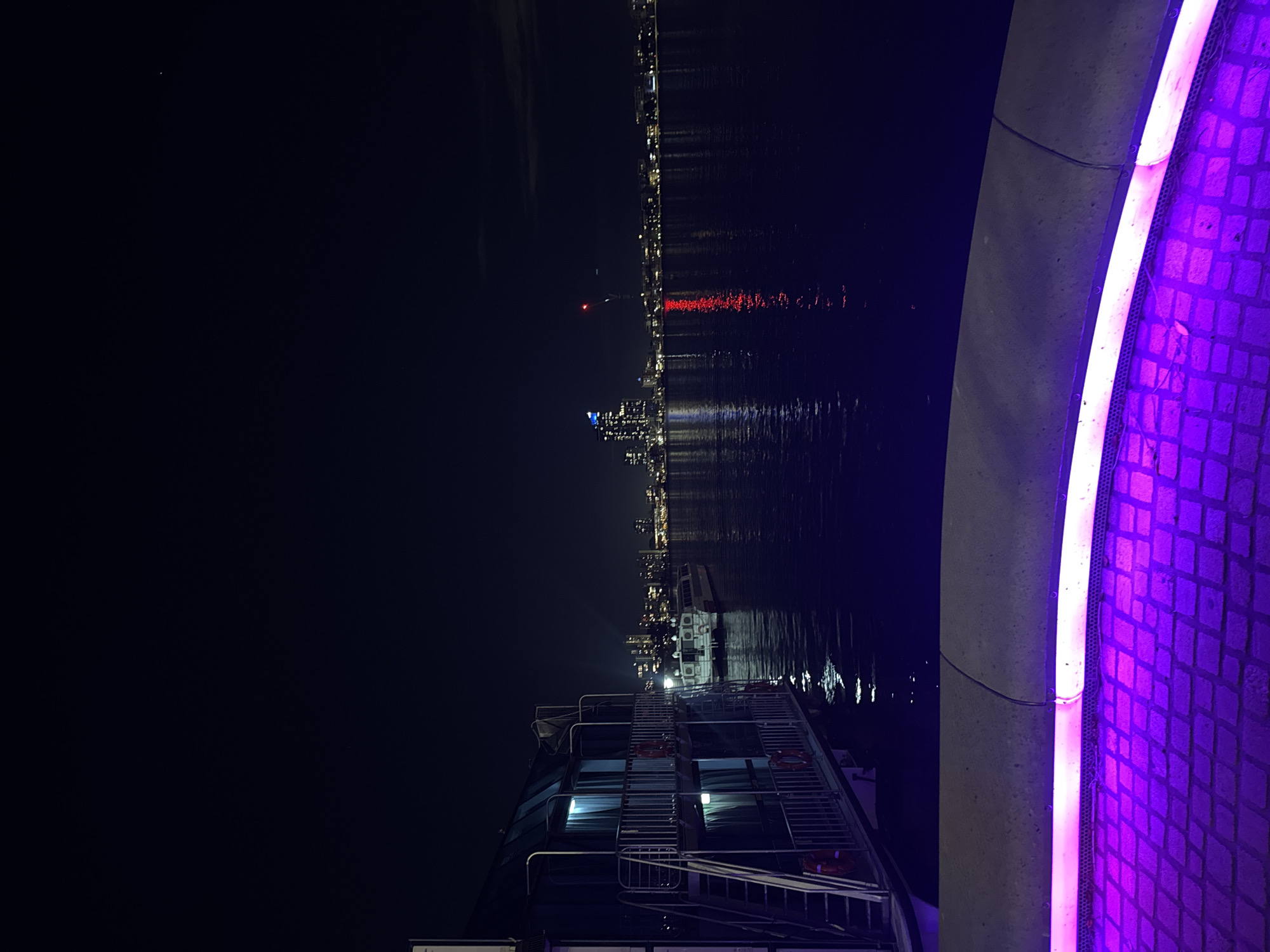 The image presents a vibrant night scene along the waterfront in Perth, Australia. The perspective is from a dock or promenade, looking across a body of water towards a distant city skyline.
In the immediate foreground, a striking curved structure, likely part of a walkway or barrier, runs along the right side of the frame. This structure features a prominent, bright purple/pink LED light strip that illuminates the textured, possibly brick or cobblestone, surface behind it, casting a strong glow.
To the left, a multi-decked boat or ferry is docked, occupying a significant portion of the mid-ground. Its interior lights are visible through the windows, and multiple railings with several orange life preservers line its decks. Stairs leading between decks are also clearly visible, suggesting it is a passenger vessel.
Beyond the boat and across the body of water, likely the Swan River, the glittering skyline of Perth stretches horizontally. Numerous buildings are illuminated, their lights reflecting brightly on the dark, calm surface of the water, creating elongated streaks of light. A particularly prominent red light, possibly a beacon or antenna on a tall structure, stands out in the distant city lights, its reflection also visible in the water. The sky above the city is dark, indicating a clear night, with a single faint point of light visible in the upper left.
No people are present in the image, and there is no visible text. The overall atmosphere is serene and vibrant, showcasing the city's illuminated waterfront after dark.