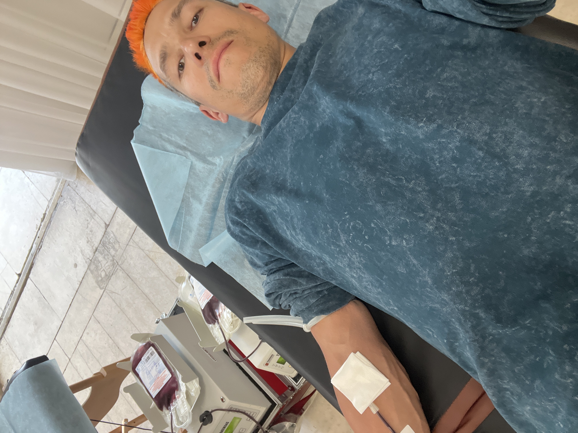 A man with orange hair is lying on a medical examination table while donating blood. His left arm has a needle inserted and is covered with a bandage. A bag of collected blood is visible next to the table, connected to the machinery used in the blood donation process. The location is presumed to be in Chaikovsky, Russia.