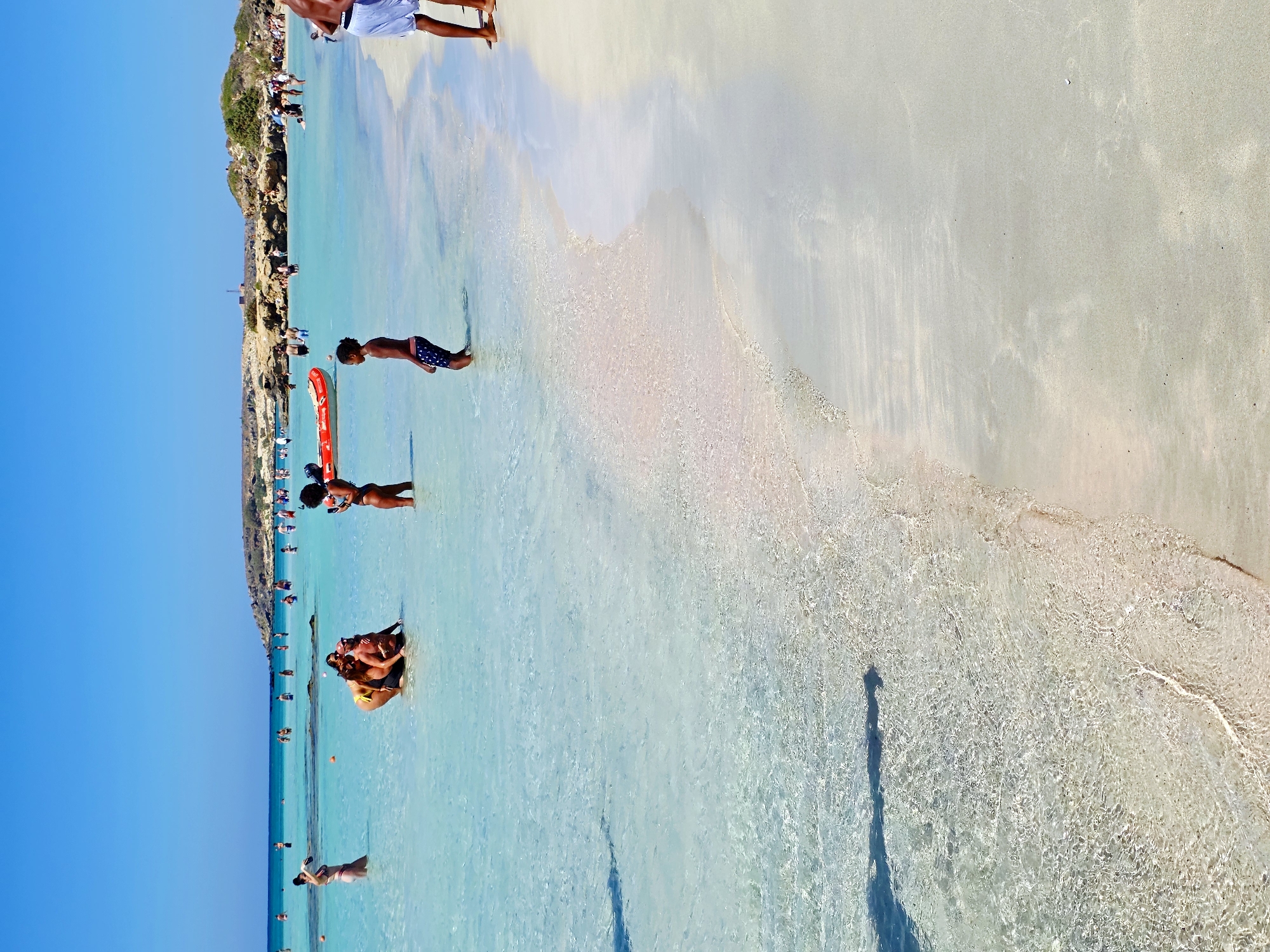 This image depicts a sun-drenched beach scene with crystal-clear turquoise water lapping at a sandy shore. Several people are enjoying the water and beach. In the foreground, a young Black boy stands in the shallow water, facing away from the camera, wearing patterned swim trunks. To the right, the legs of a person wearing white shorts are visible, standing on the dry sand.
Further out in the water, a group of individuals are interacting. Two women, one in a yellow bikini and the other in a patterned bikini, are posing closely together in the water. Another woman in a bikini is taking a picture of them with her phone. Further back, a red inflatable raft is partially visible. Numerous other beachgoers are scattered throughout the water and on the shore in the mid-ground and background. The coastline features rocky formations and vegetation in the distance under a clear blue sky. The water is exceptionally clear, allowing visibility of the sandy bottom. The sand exhibits a light, possibly pinkish hue near the water's edge, transitioning to a paler tone further up the beach. The overall impression is one of a vibrant and pleasant summer day at a popular beach destination. The location is identified as Unknown, Greece.