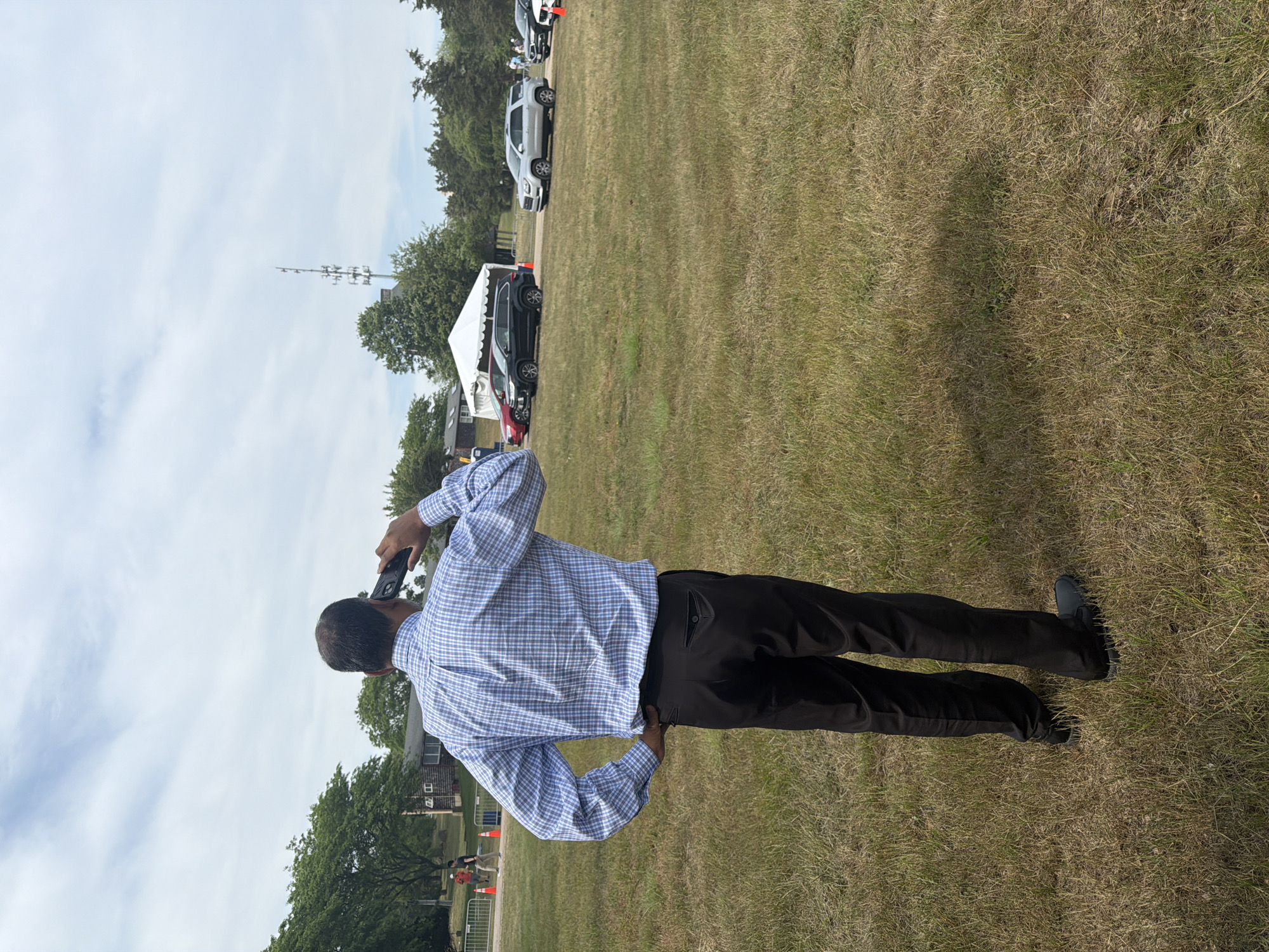 A man in a blue and white checkered button-down shirt and black pants stands in a dry, grassy field, talking on a cell phone held to his right ear. His left hand is on his hip. In the background, there are several vehicles, including a black SUV and a silver SUV, parked in a line. A white tent is set up near the vehicles. Trees and a cell tower are visible in the distance. The sky is partly cloudy. The location is identified as the Town of Southampton, United States of America.