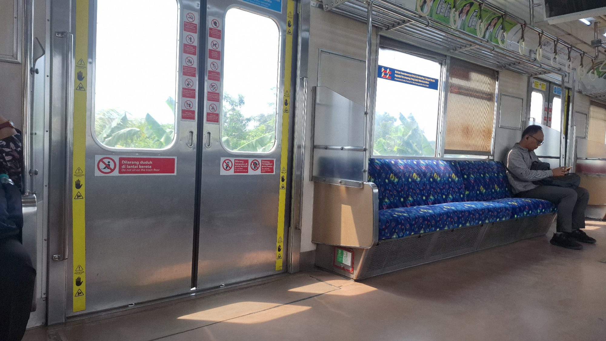 The interior of a train car in Tambun, Indonesia, is captured in the image. A seated Asian man wearing glasses and a long-sleeved shirt is looking at his phone while a black bag is in his lap. The man is sitting on a blue patterned seat with metal supports. A closed metal door with a yellow handhold strip is on the left side of the image. A red sign on the door reads "Dilarang duduk di lantai kereta. Do not sit on the train floor." Above the door, advertisements are attached to metal racks. There is a window with a brown blind, and outside the window, trees and sky are visible. The car floor is a light brown color.