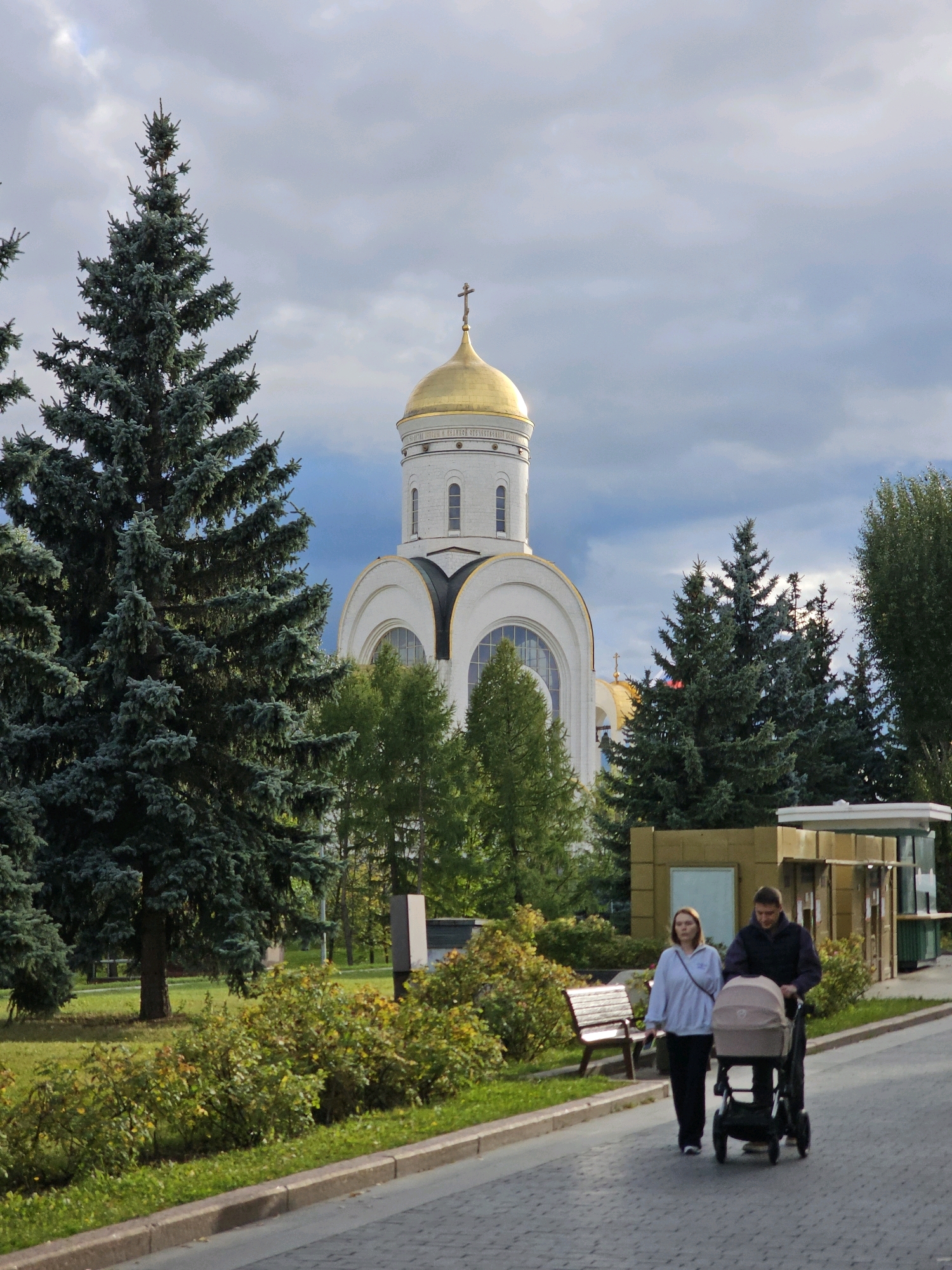 This is a medium shot of a couple pushing a stroller on a paved path in front of a white domed building with a golden dome. The building appears to be a church, and the golden dome is topped with a cross. The couple consists of a woman in a light-colored hoodie and black pants, and a man in a dark jacket and pants. The stroller is tan and has a black frame. The path is lined with grass and bushes. In the background, there are several tall pine trees and a cloudy sky. The scene is likely set in a park or public space in Moscow, Russia, given the architectural style of the building and the presence of pine trees. The overall impression is one of a serene and pleasant day.